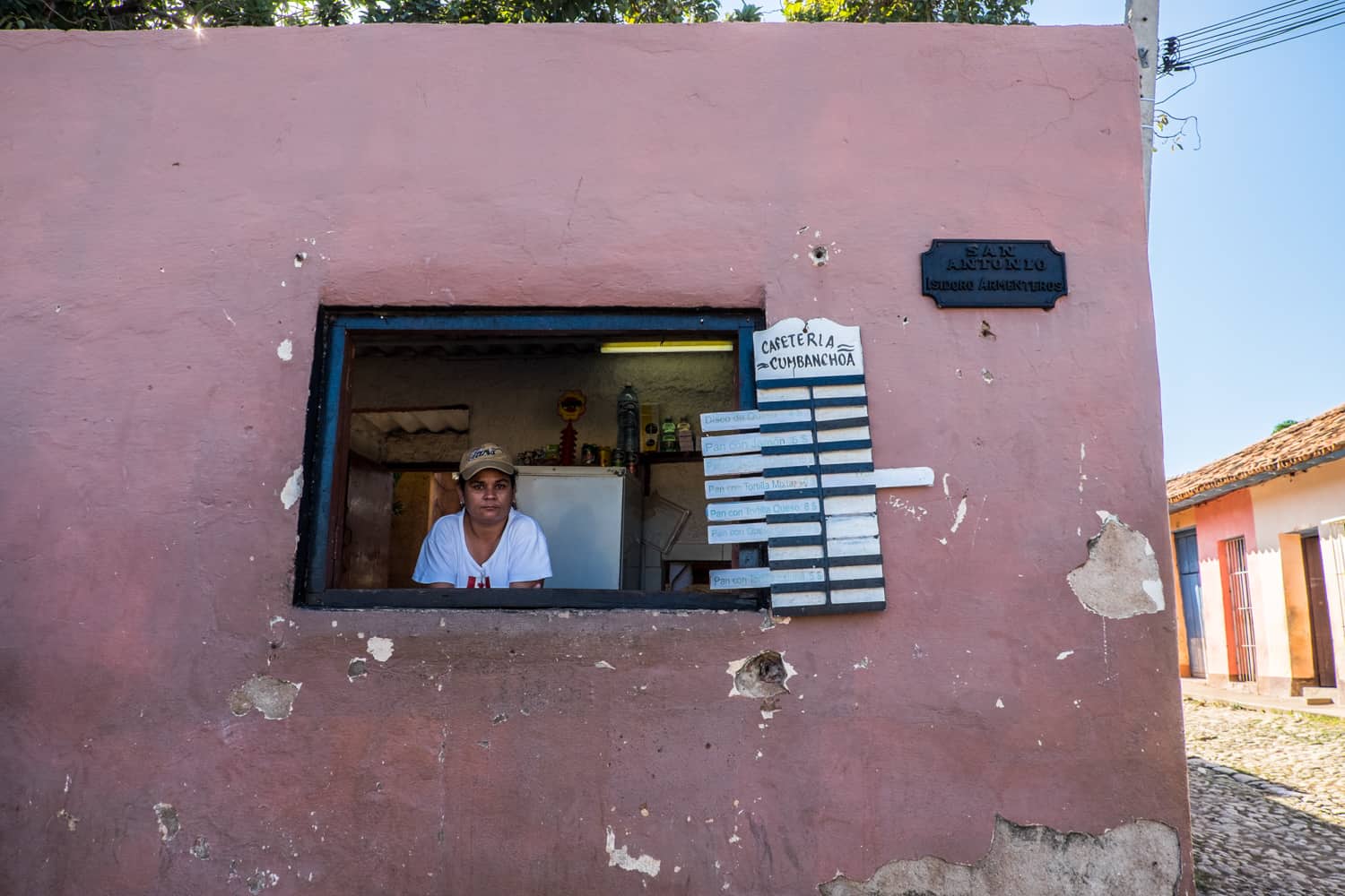 A woman at a window on a pink painted building with a Cafeteria sign listing food and drinks to buy. 