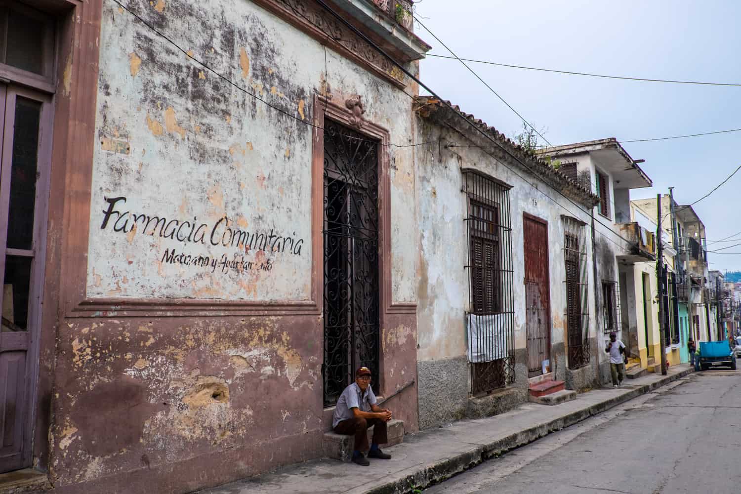 A man sits on the window ledge of a peeling brown and white building that has the words Farmacia Comunitaria painted on it. 