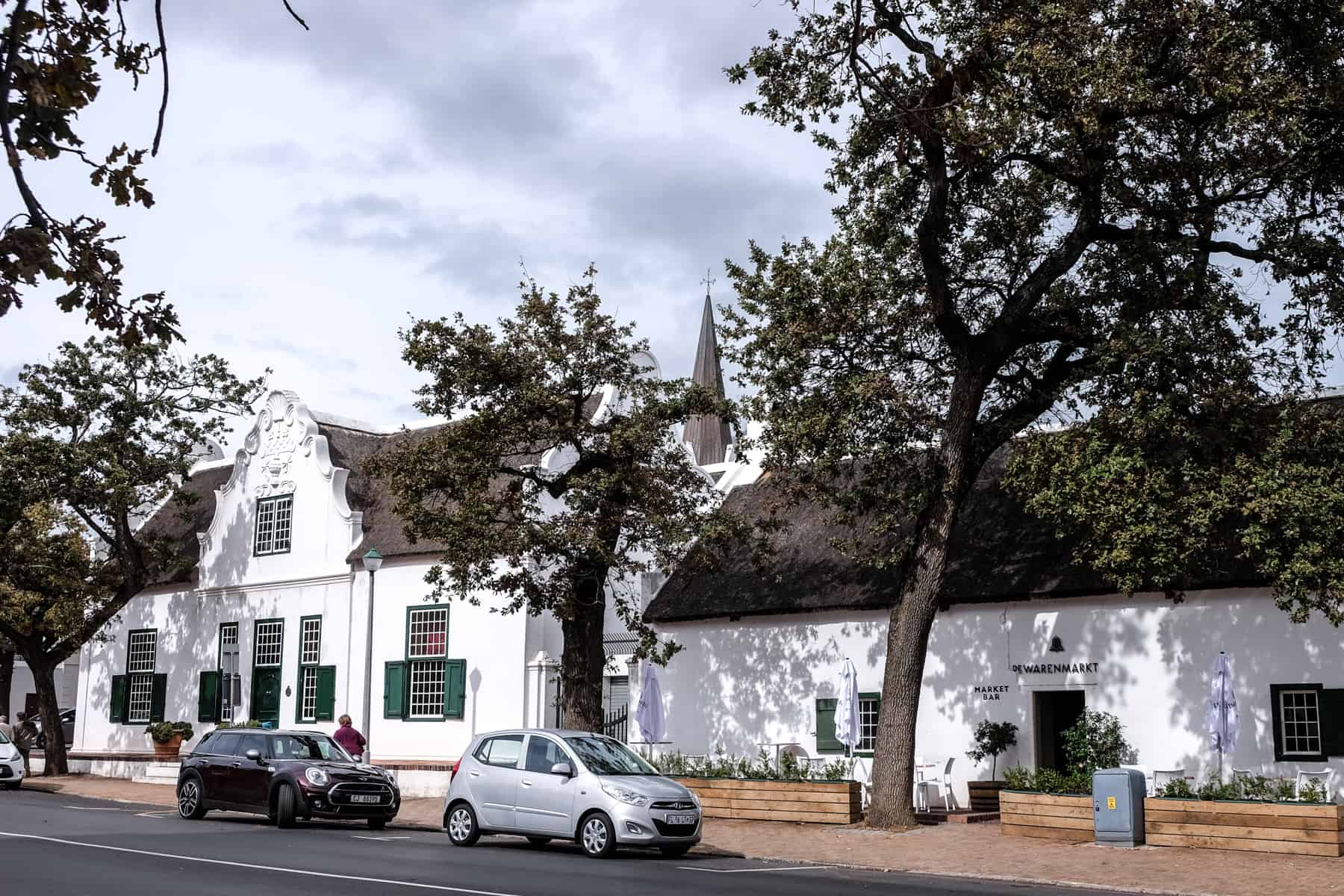 A long white classic-style building with a dark brown thatched roof surrounded by trees - the exterior of the De Warenmarkt market bar in Stellenbosch 