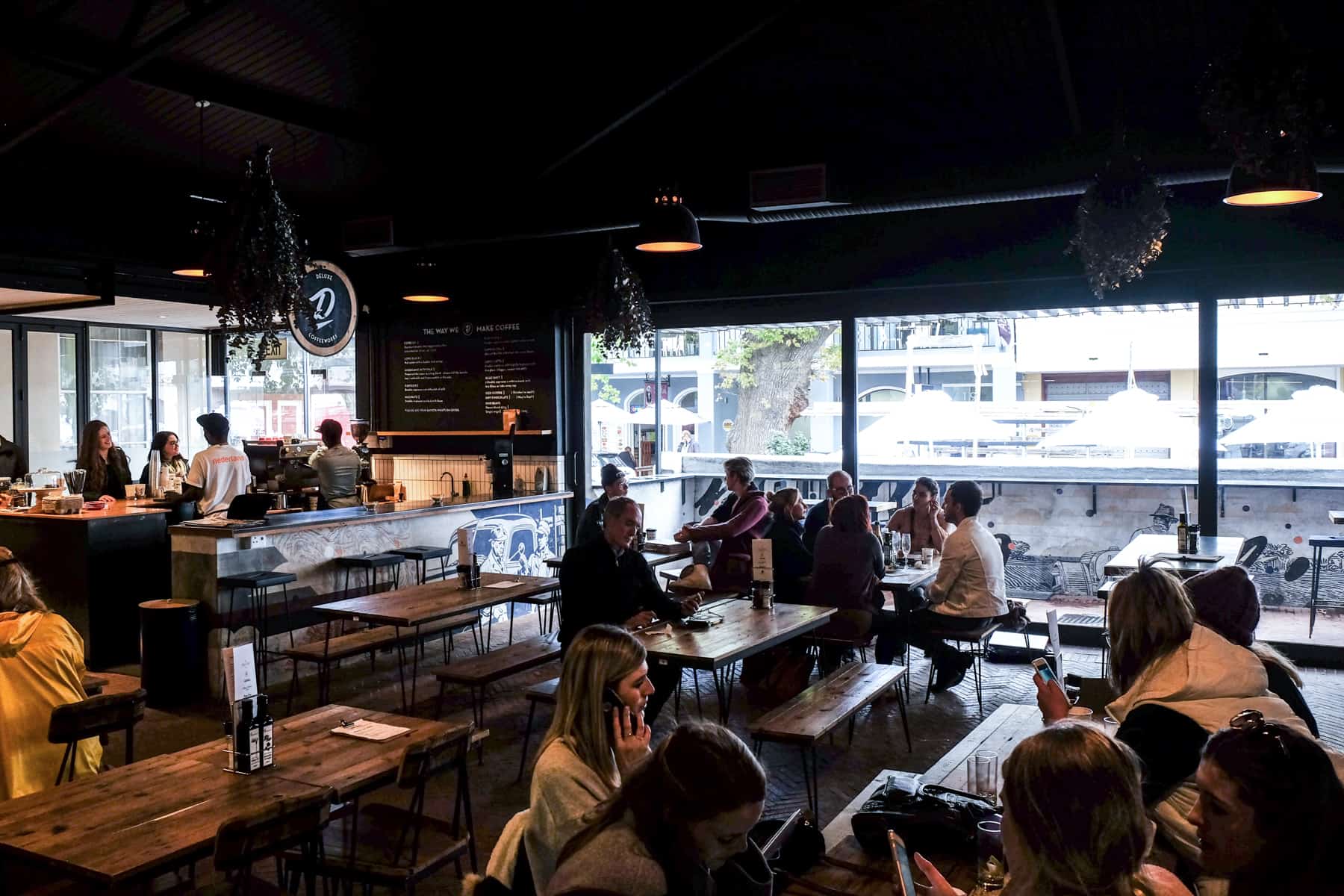 People sit at wooden tables inside the dark interior of the De Warenmarkt restaurant in Stellenbosch. Large glass windows provide the light against the almost black ceiling 