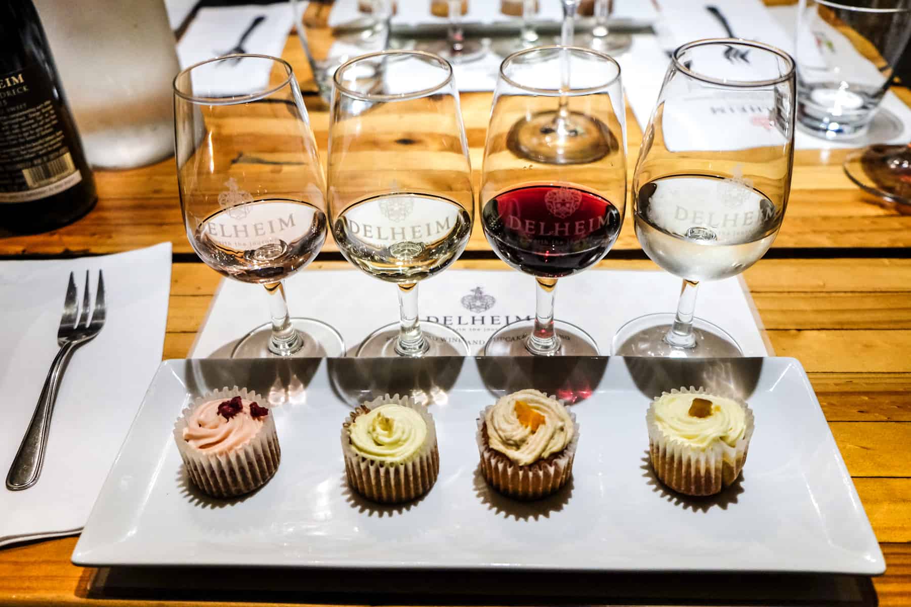 Four wine glasses (three white and one red) lined up behind four cupcakes - part of the Delheim Wine Estate wine and cupcake pairing experience in Stellenbosch, South Africa 