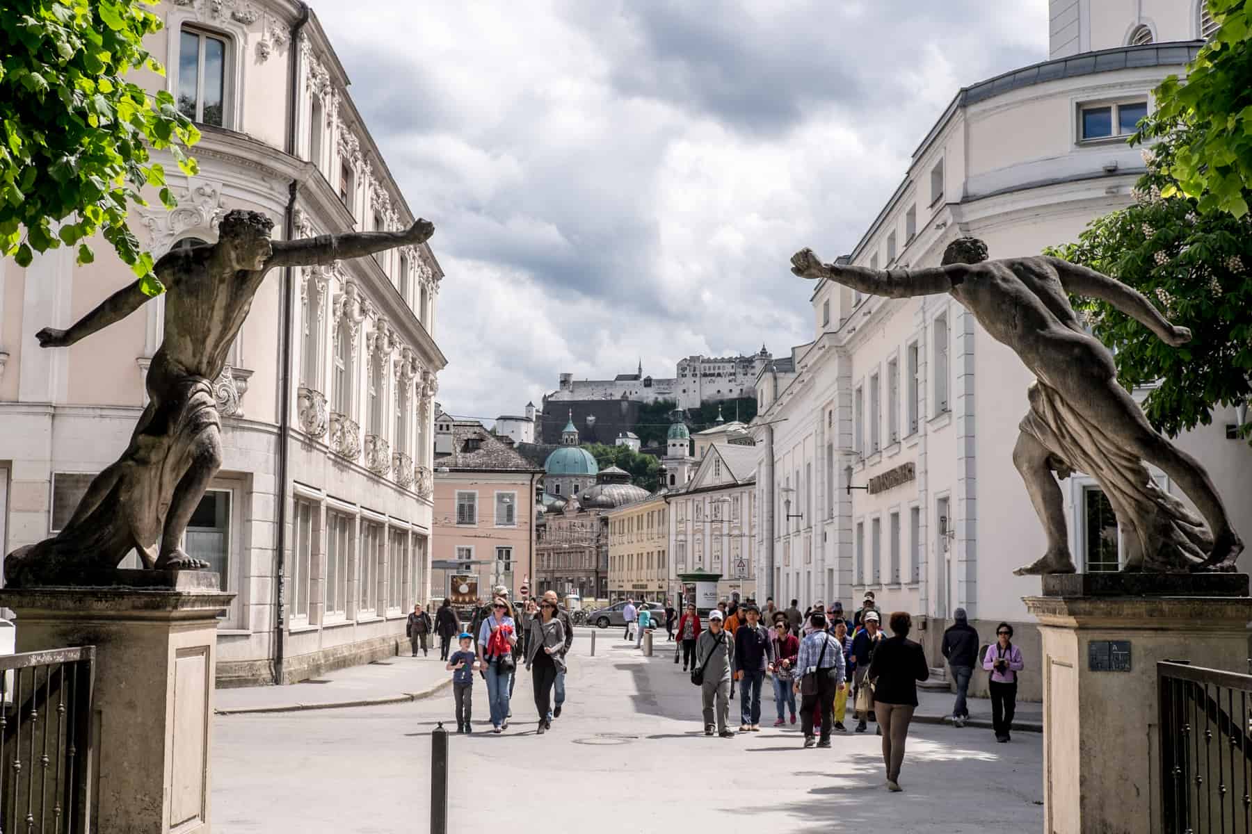 Two tall statues with arms outstretched are the decor on the Gate of the Mirabell Gardens in Salzburg, Austria. In the distance is the hilltop fortress.