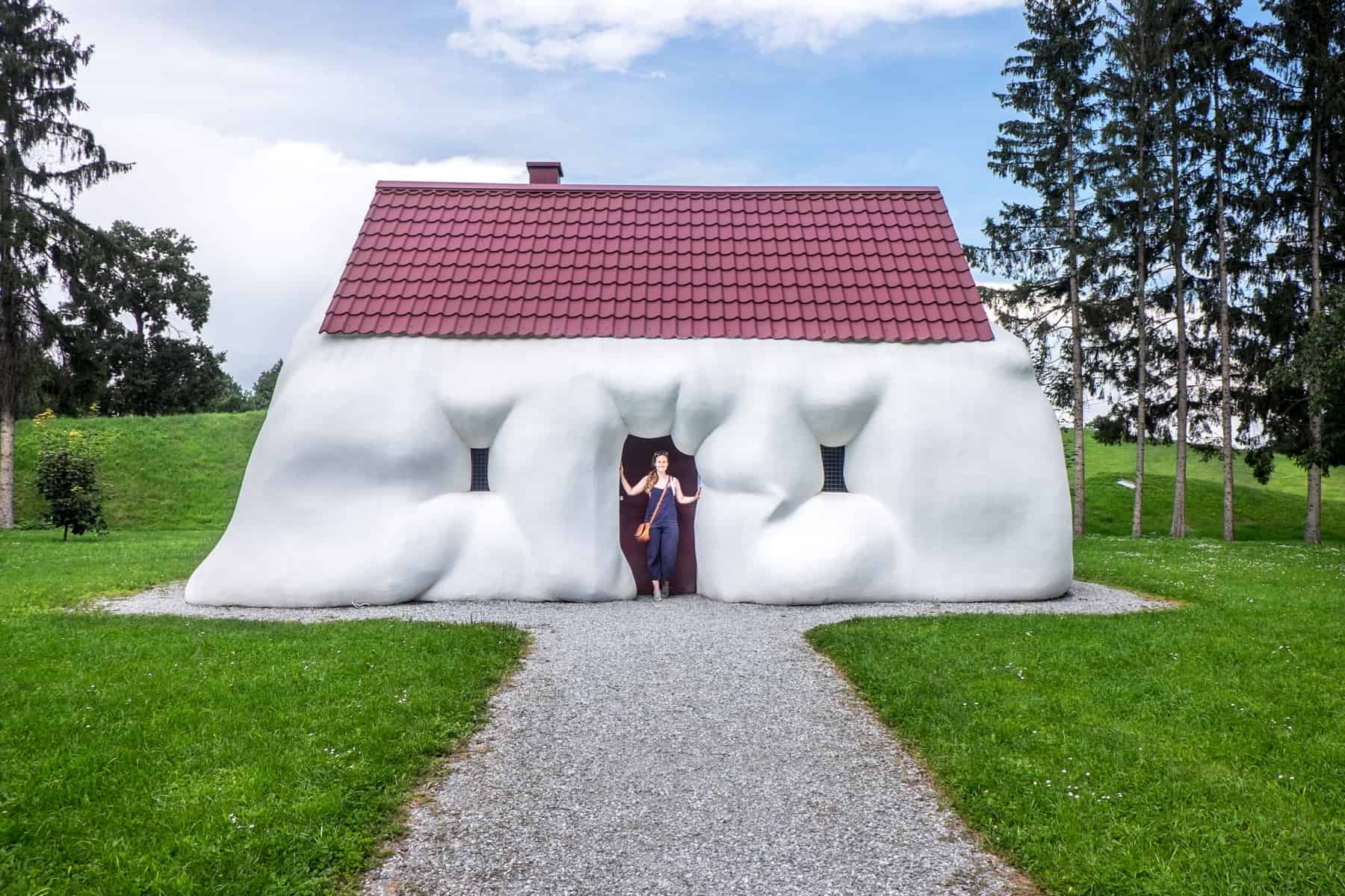 A woman stands in the doorway of the 'Fat House' artwork, a white house with a red roof that looks like its almost melting. Part of the Austrian Sculpture Park on a day trip from Graz