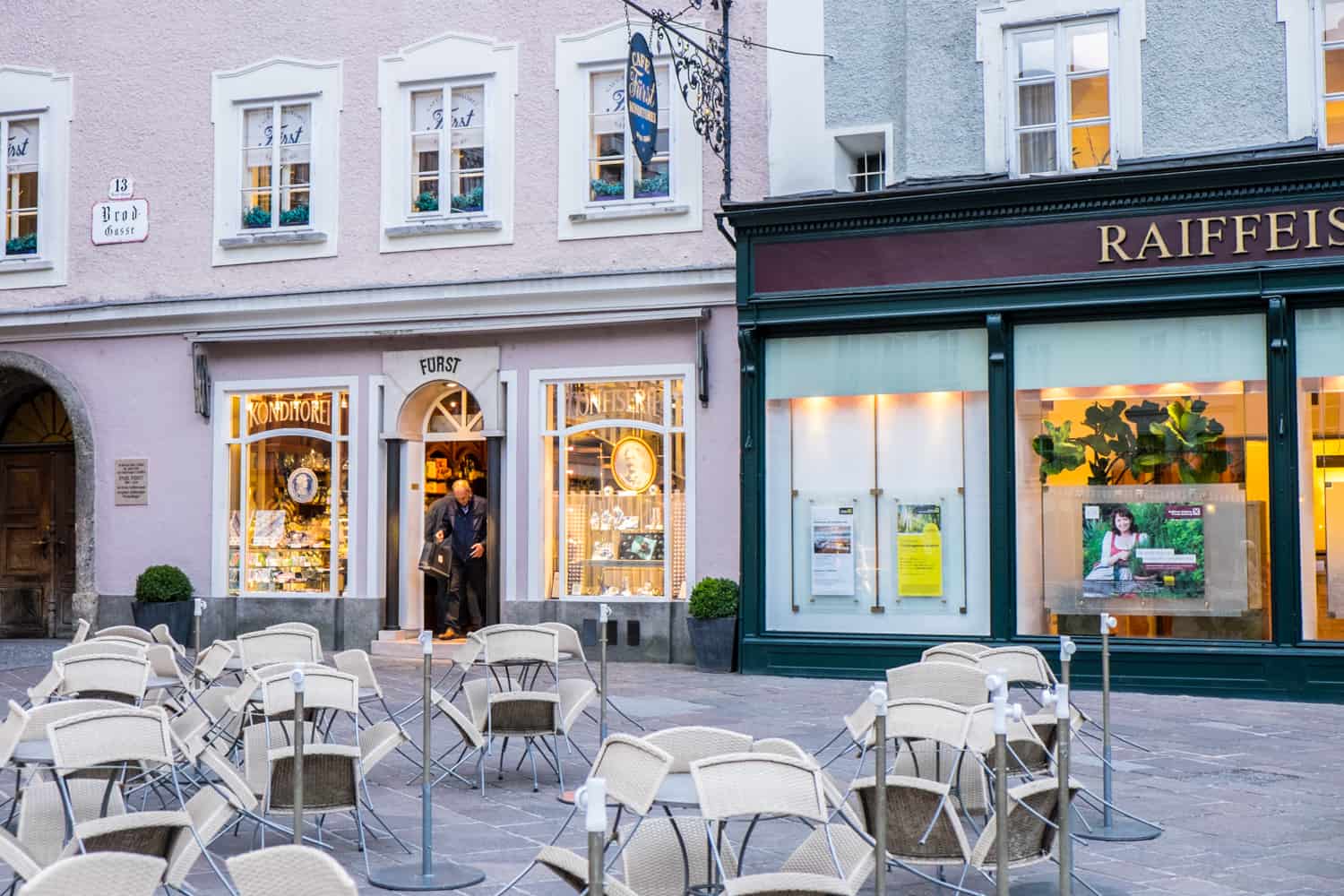 White chairs and tables outside a pink building with white window trims, golden lights and the name over the door arch: "Fürst" - a famous chocolate shop in Salzburg. 