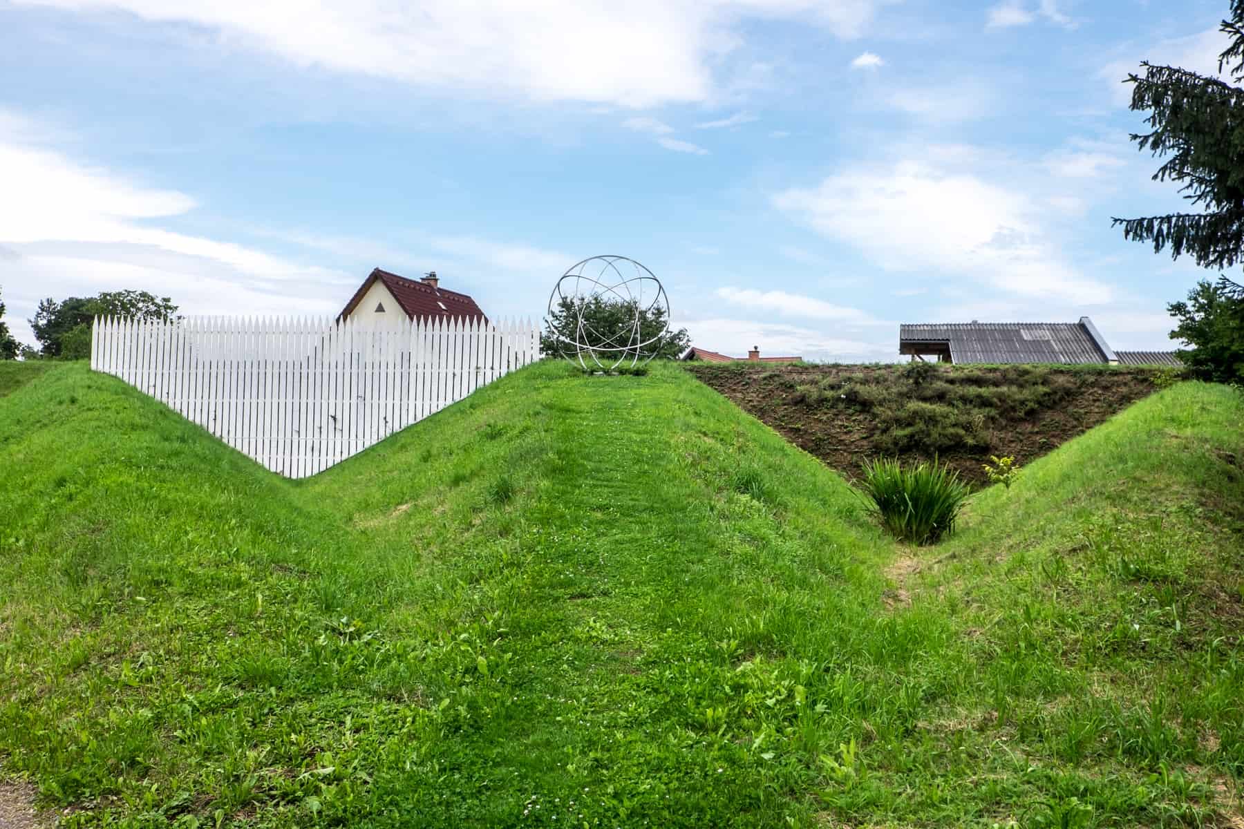 A house with a white fence and a metal artwork sit on top of three triangular green mounds at the Austrian Sculpture Park near Graz
