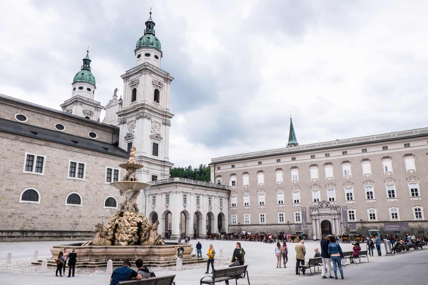A Public Square with beige and white building with mint green spires surround a Golden Fountain in Salzburg.