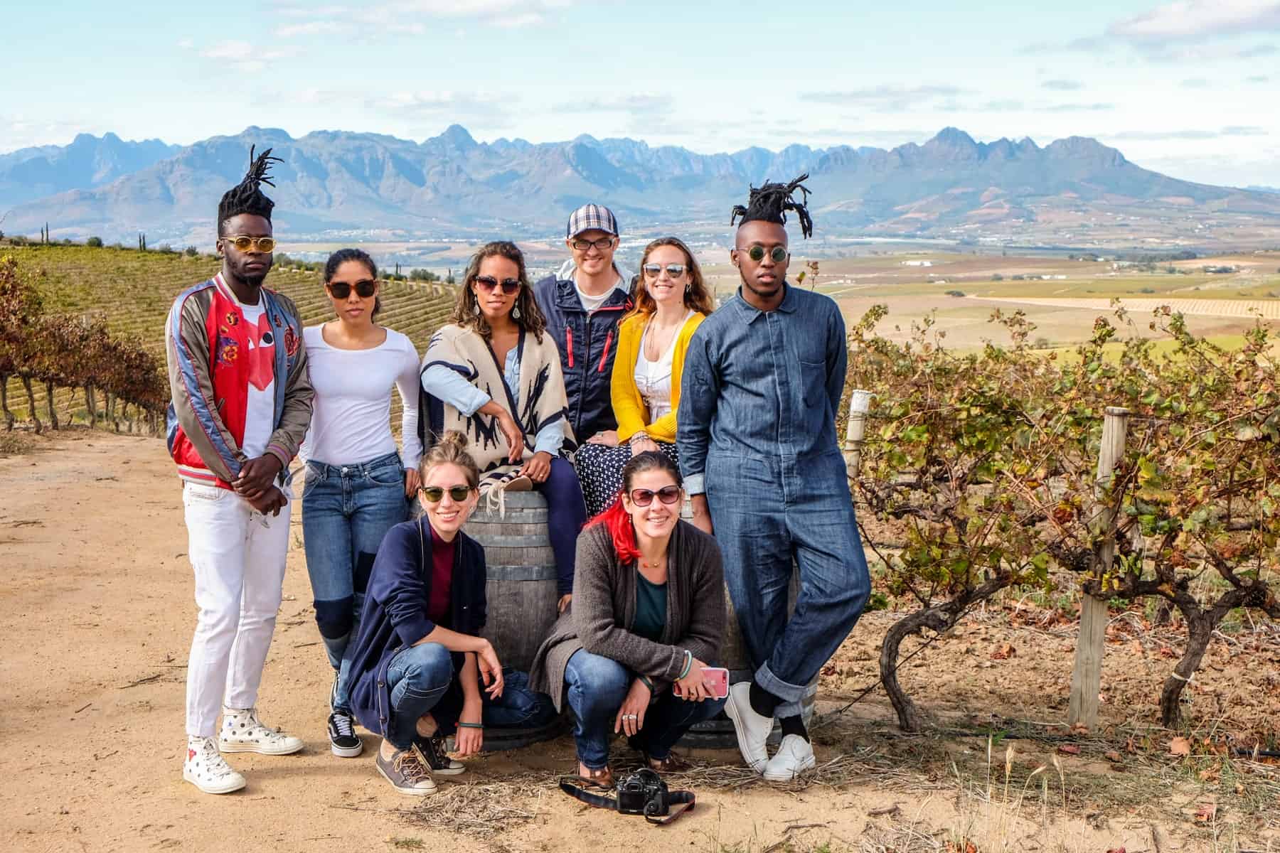 A group of 8 people pose with a wine barrel on an orange dirt road in the wide, open, flat lands of Jordan Wine Estate in Stellenbosch