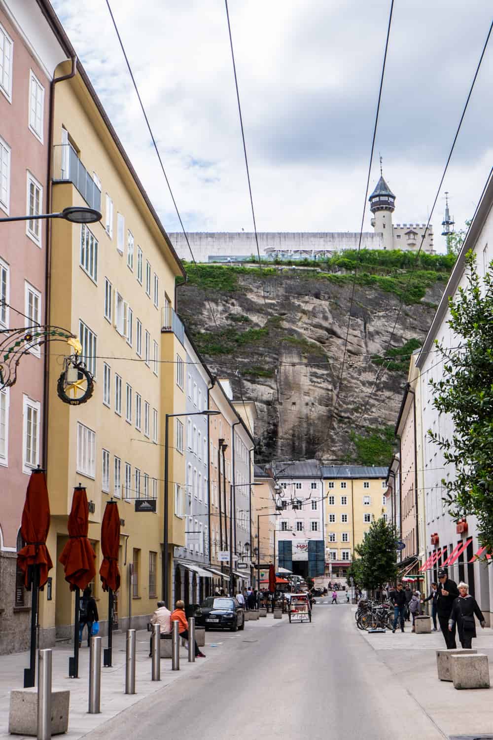 View down a long street lines with pastel coloured buildings and which looks up to a large rock wall with a white fortress on top. 