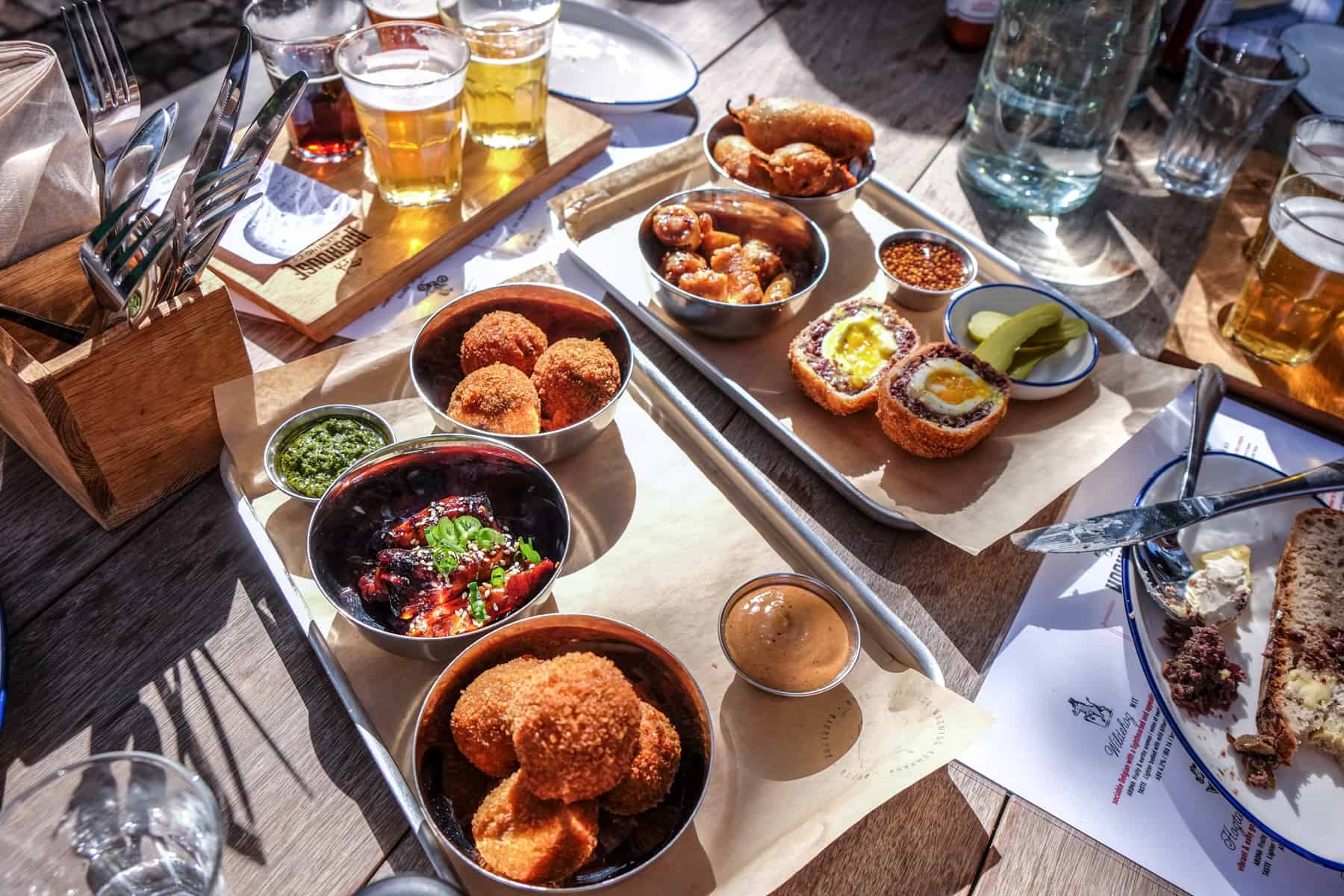 Two silver trays contained breadcrumbled balls, bowls of green and red sauces and a cut pork pie. Behind the trays is a tasting platter of four beers. The selection is from the Hoghouse BBQ at Spier Wine Estate 