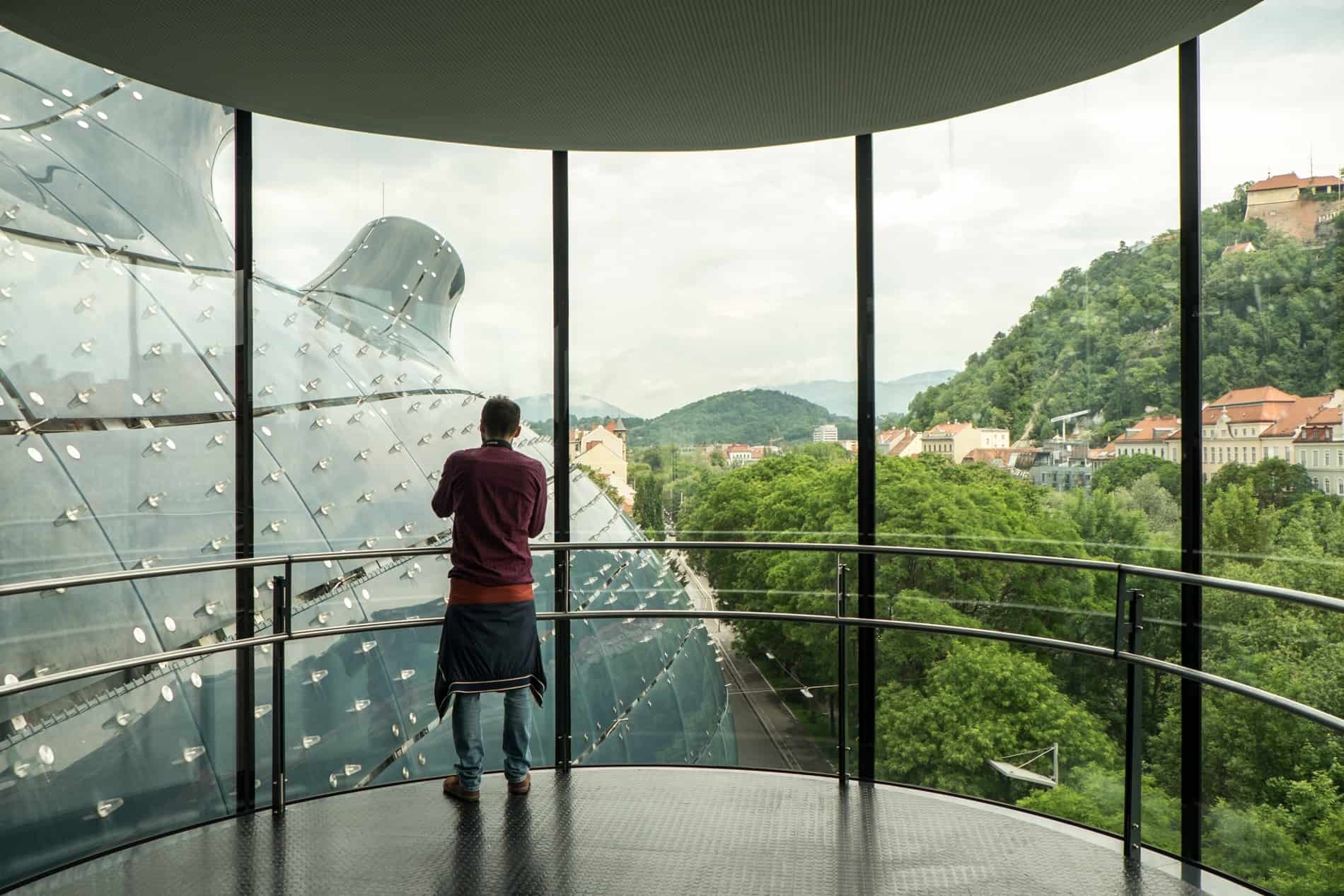 A man stands at a floor-to-ceiling window overlooking a blue, boubous structure, from inside the Kunsthaus modern art musuem in Graz. 