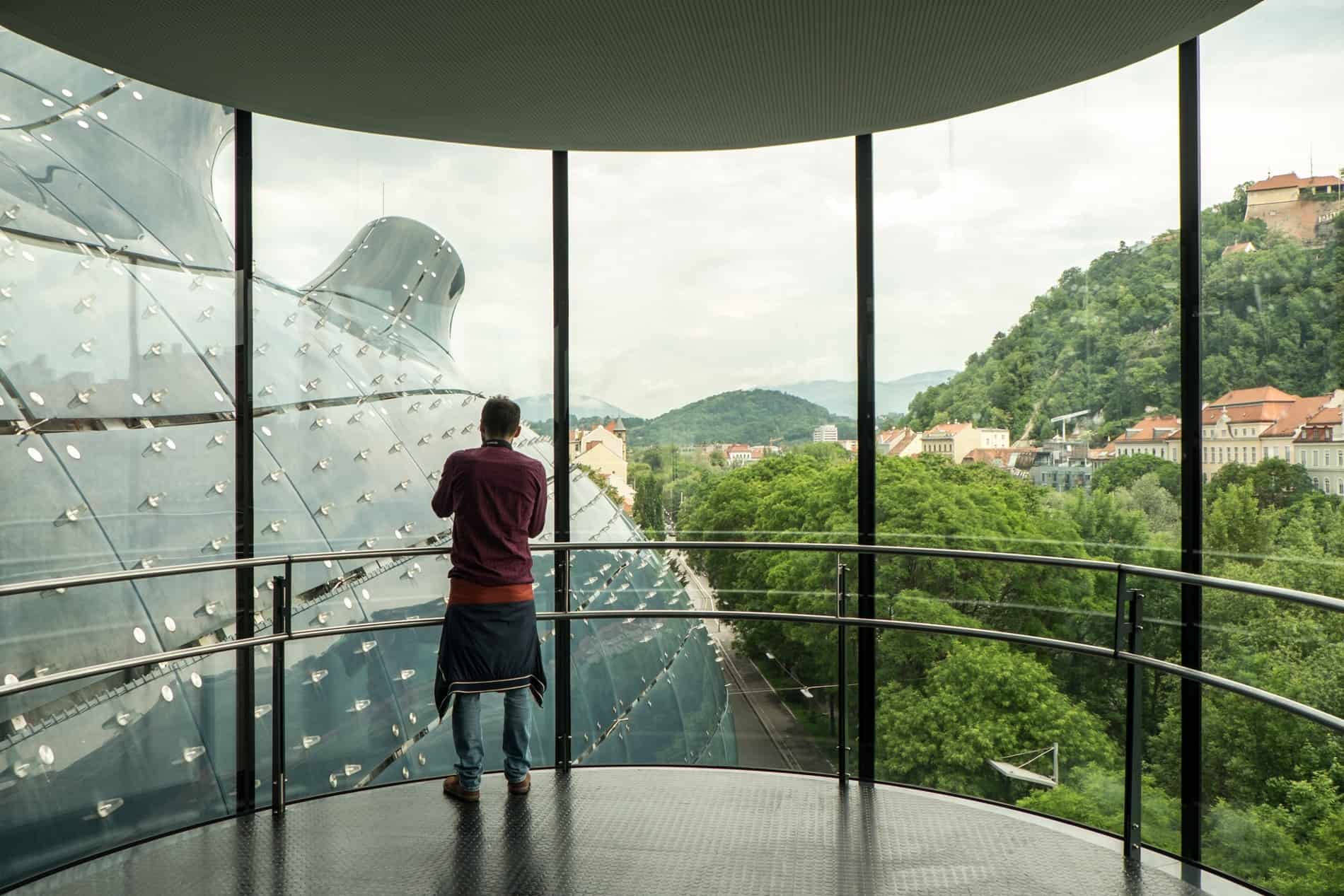A man stands at a floor-to-ceiling window overlooking a blue, boubous structure, from inside the Kunsthaus modern art musuem in Graz.