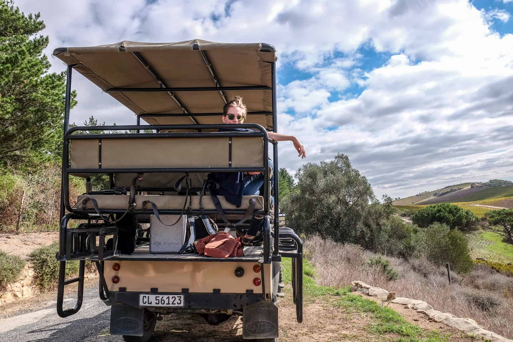 A woman wearing sunglasses looks out over the top of a backseat of a jeep, while her arm hangs over the side of it. The jeep is on a gravel road with green hills in the distance