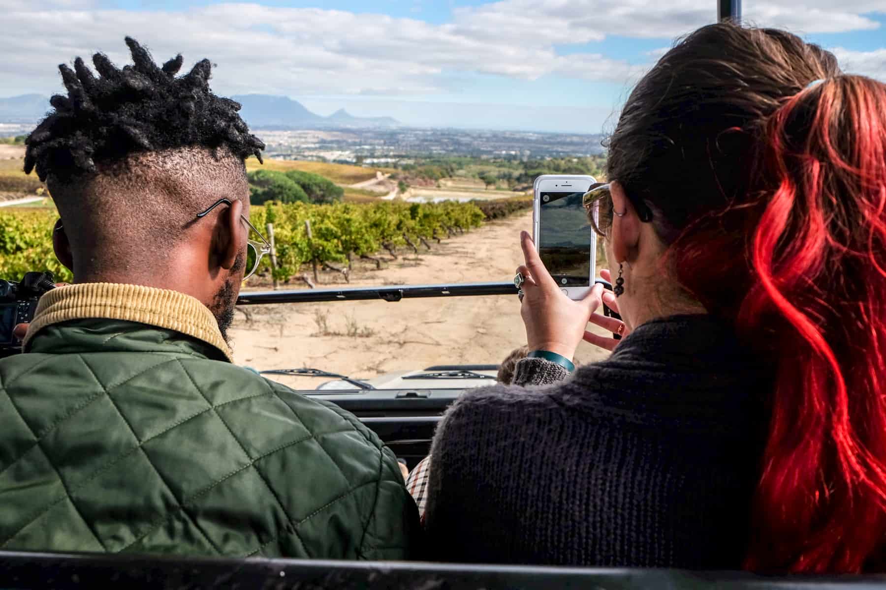 A woman with red hair takes a picture of the landscape before her on her mobile phone while on a jeep. She sits next to a man looking straight ahead at the view