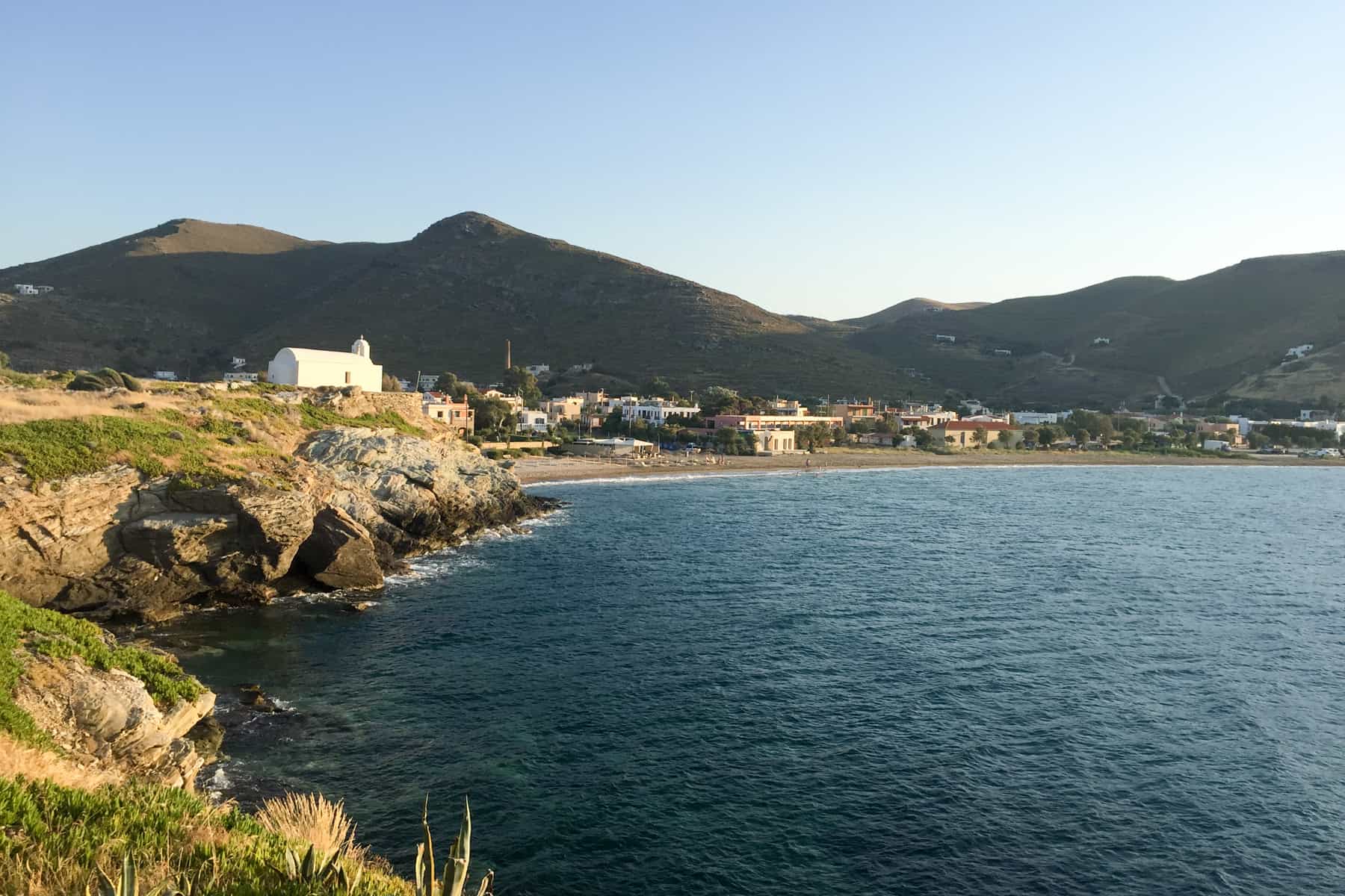 A sunset glow on a coastal cove on Kea Island. The water is a dark blue, the land is full of rugged grass and a white house pokes through it in the distance