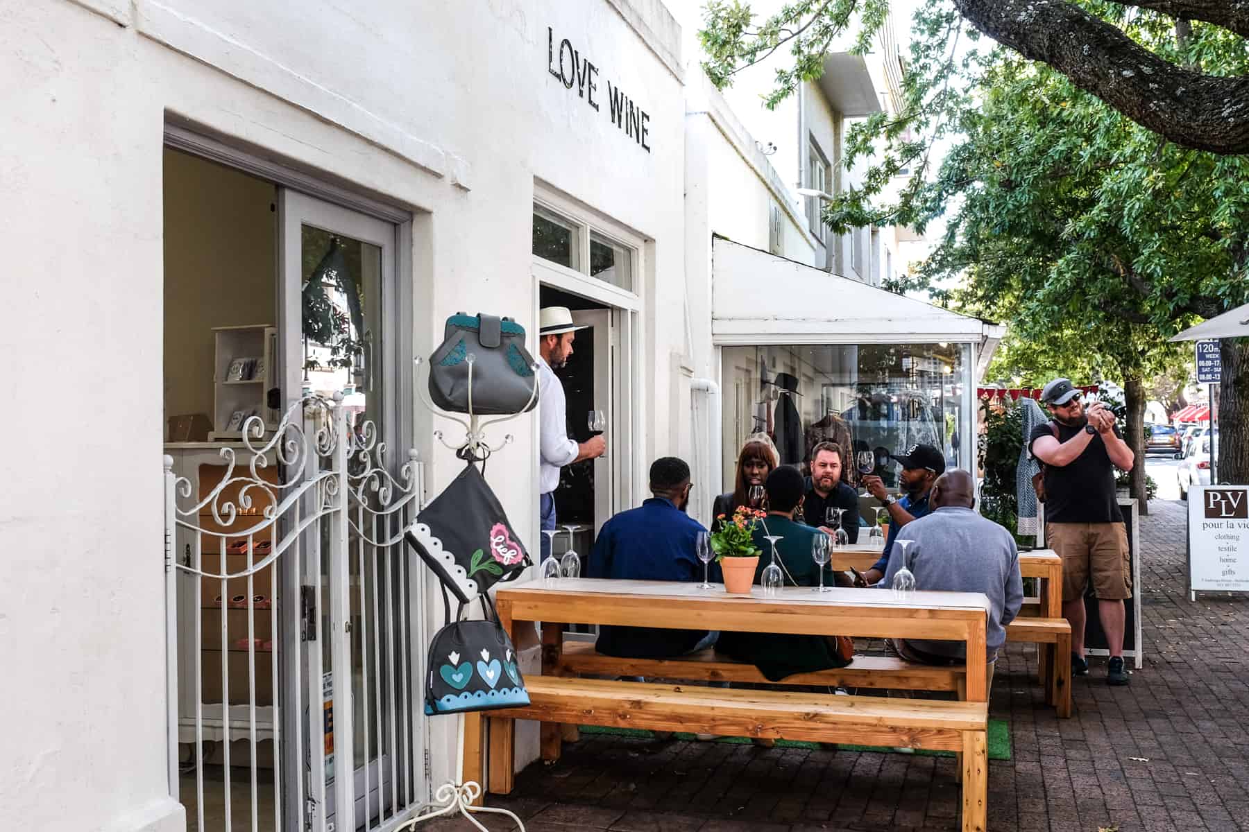 A small group of people site on a golden wooden bench outside a white building in Stellenbosch. The wording above them on the wall reads 'Love Wine'