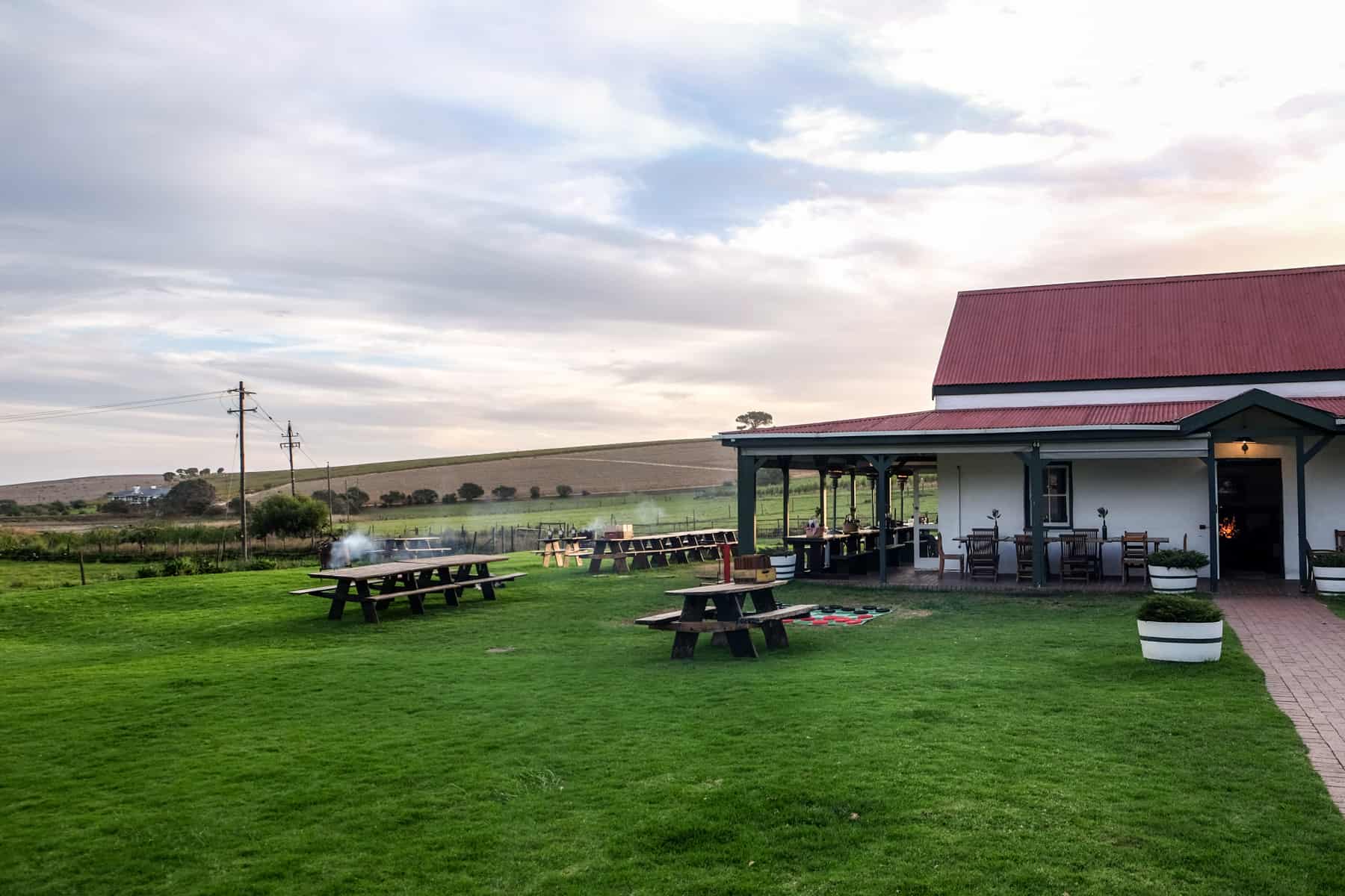 A white house with a red roof stands within a wide green field with brown seat benches and tables - part of the Middelvlei Wine Estate in Stellenbosch