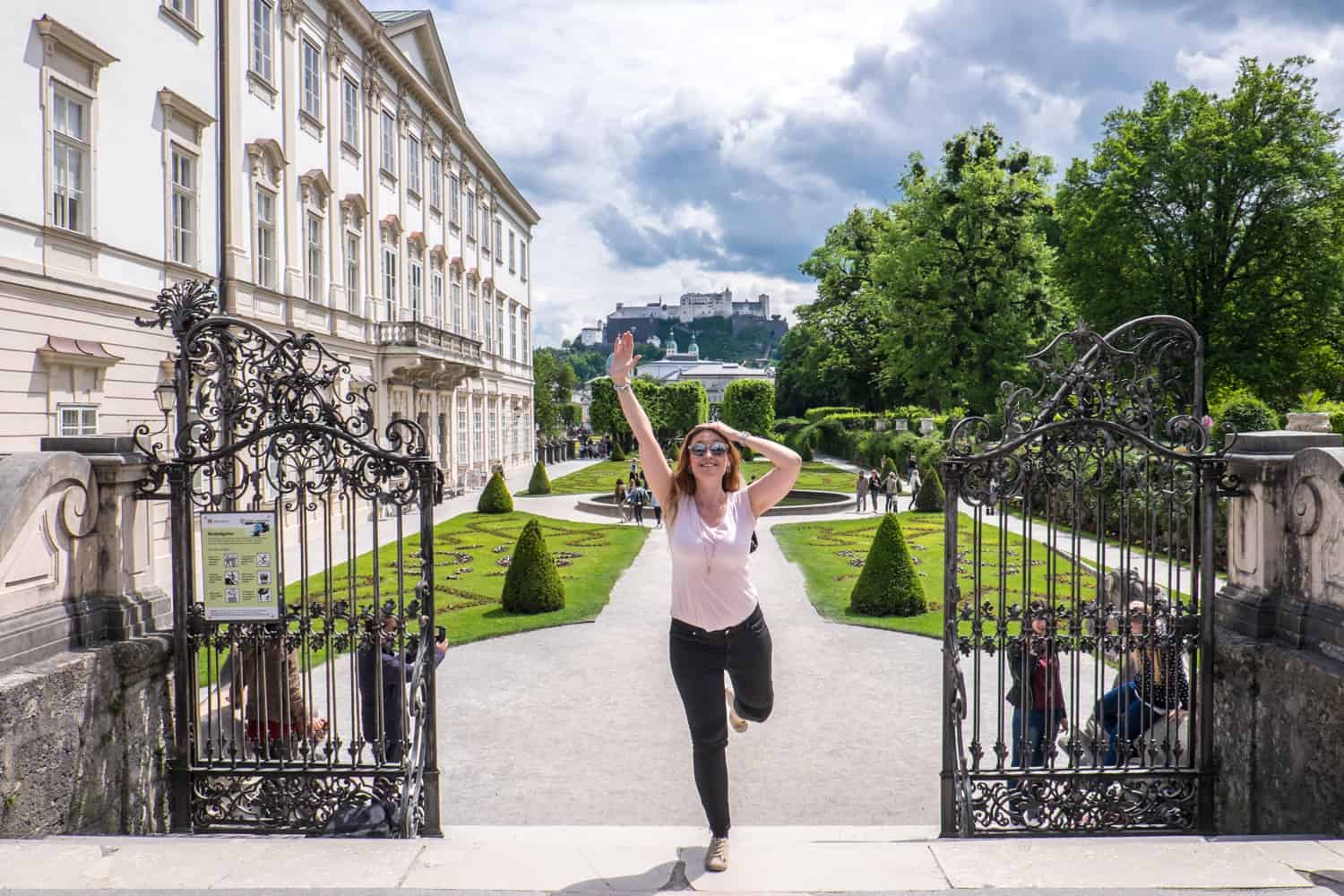 A woman in black jeans and a pink t-shirt stands at the top of the stairs, at the black gated entrance to a palace garden, with her arms raised. Behind her is a manicured garden with a fountain, and a raised fortress complex. 