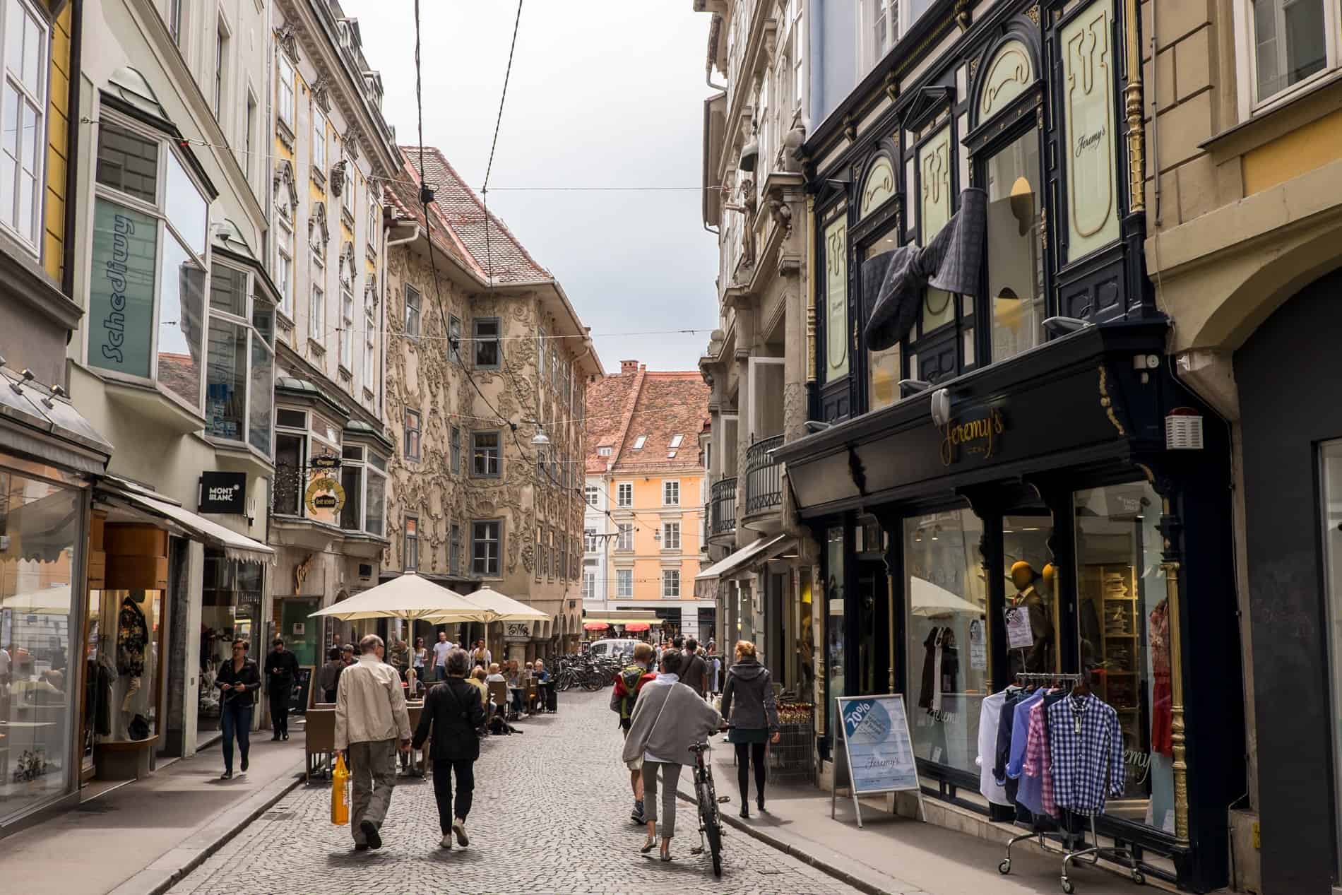 People walking down the beautiful, old Sporgasse street in Graz lined with decorated facades and exquisite shopfronts. 