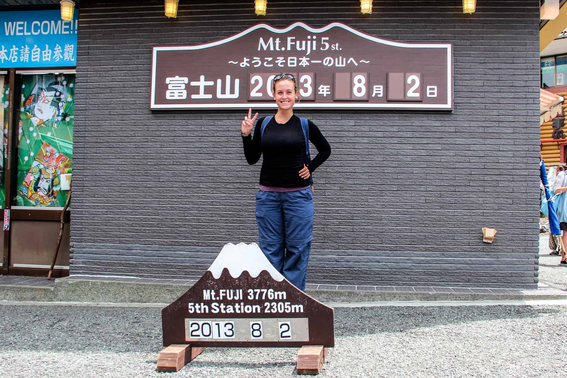 A woman smiles to the camera and makes a peace hand gesture at the sign which reads 'Mt. Fuji 3776m. 5th Station 2305m' and the date 2nd August, 2013.
