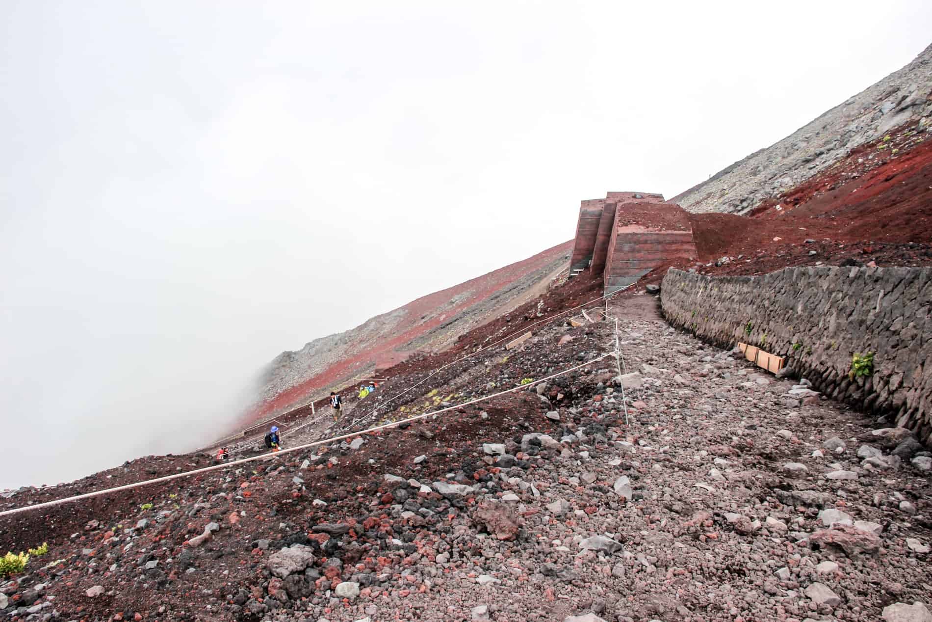 People climbing Mount Fuji on a very steep and loose rock path to a backdrop of mist. 