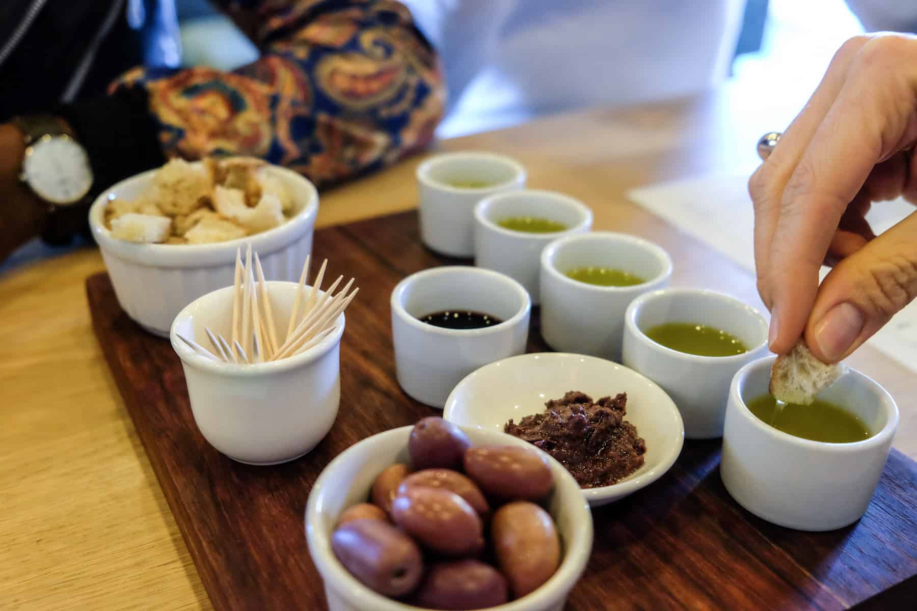 A hand dips a tiny piece of bread into one of five small white pots containing olive oil for tasting. Also on the dark wooden plate are bowls of olives and toothpicks