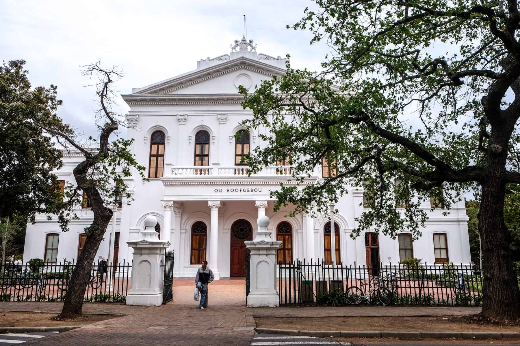 A white columned building with a triangular roof in Stellenbosch, set behind a small black fences and 2 large trees 