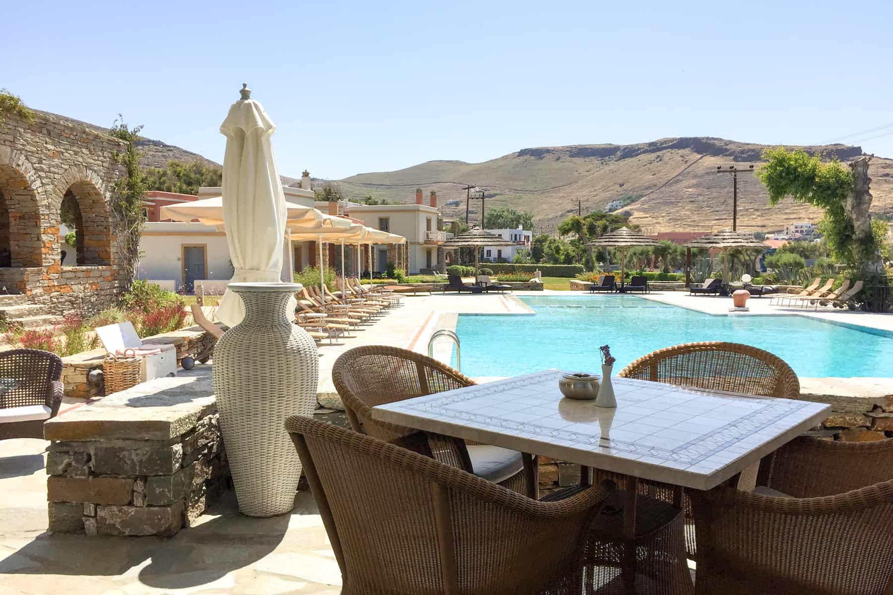 A four seater table next to a bright aqua swimming pool at Porto kea Suites in Kea, Greece backed by low rugged yellow-green hills