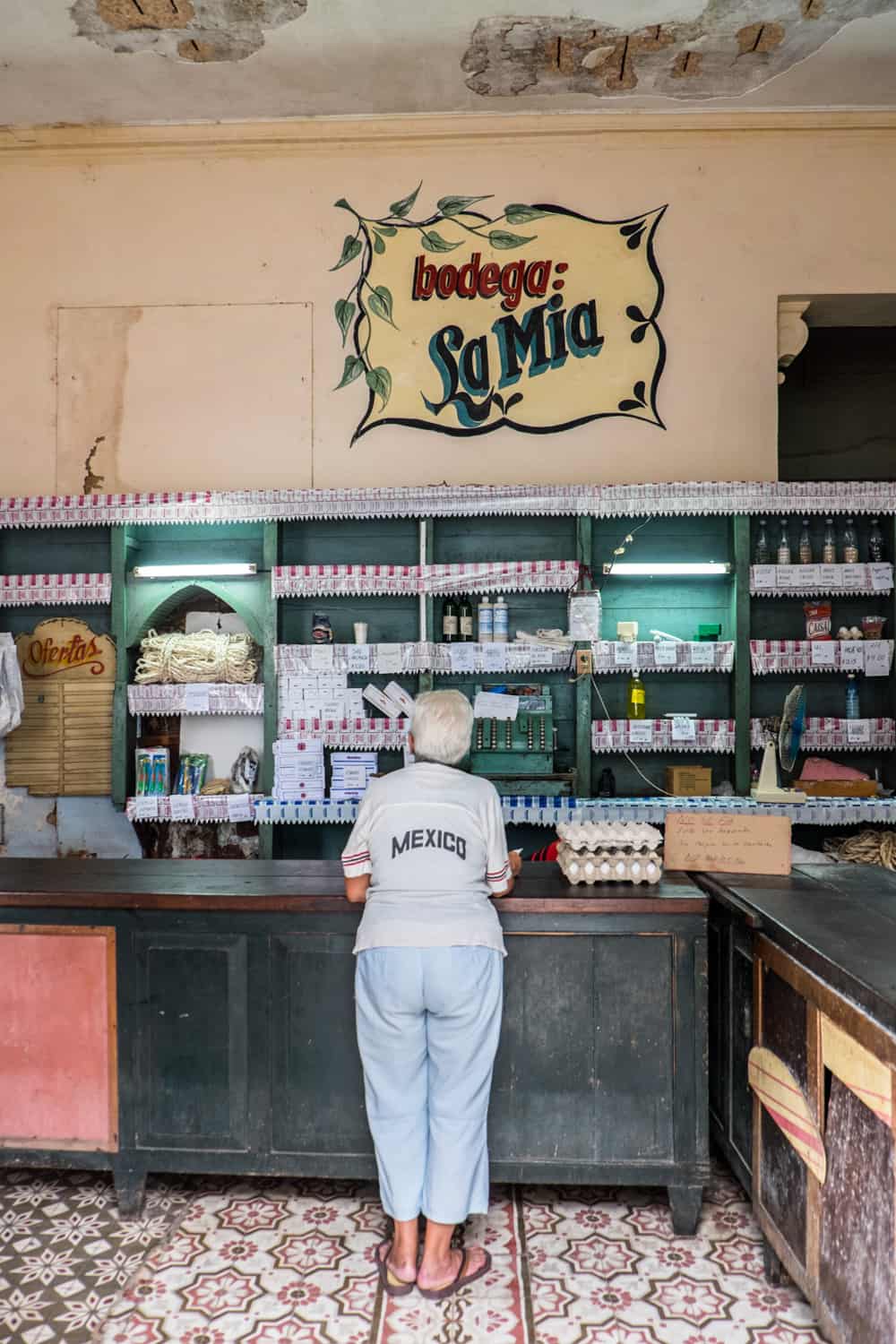 A woman in a Mexico t-shirt stands at the counter of a ration store in Cuba next to two trays of eggs.