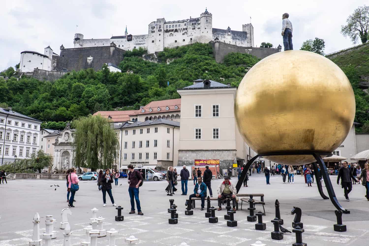 Modern artworks in Salzburg include a man standing on a giant golden globe and a large interactive chessboard. Both are found in a public square below a hilltop fortress. 