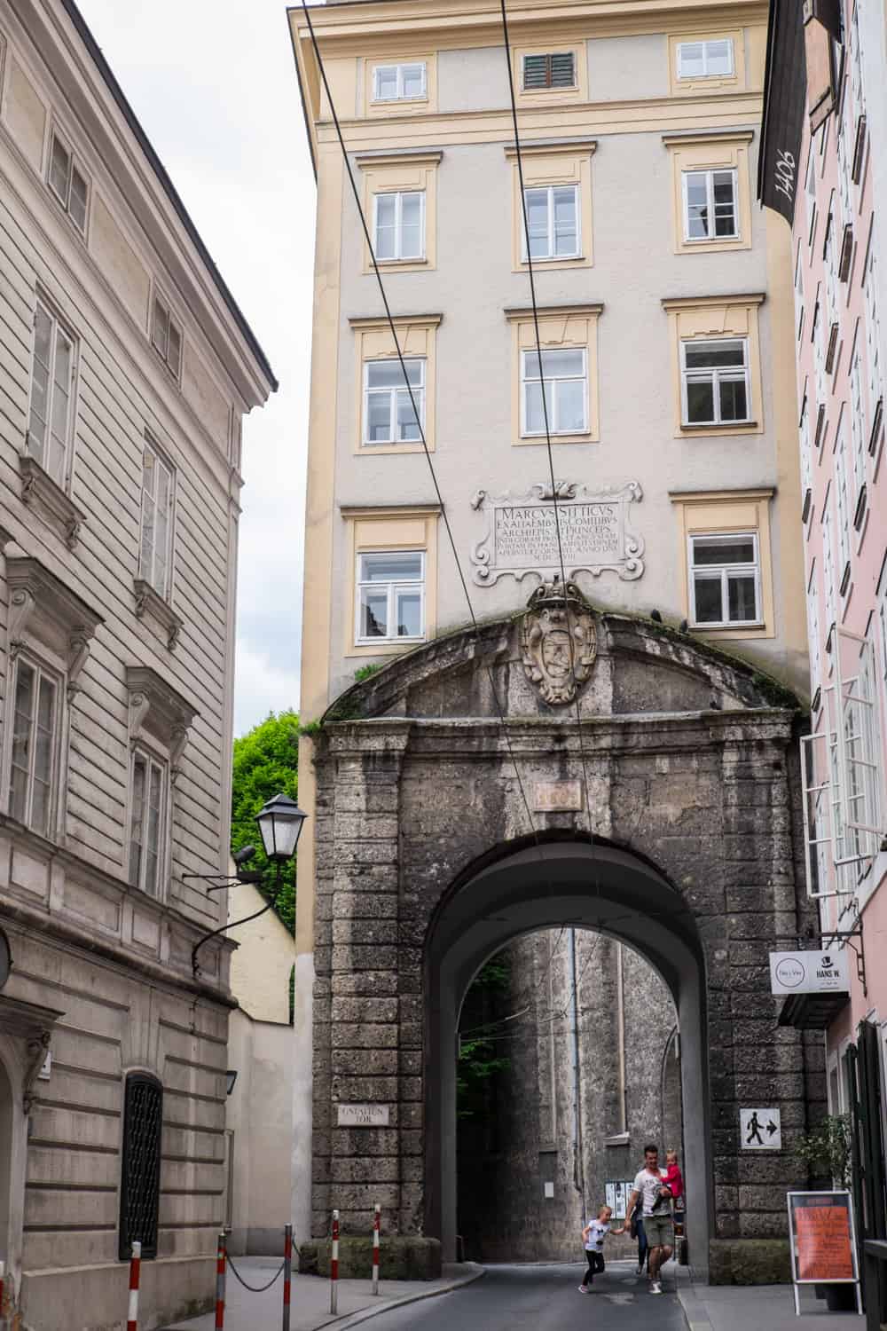 A darken cream stone old city gate in Salzburg with a modern yellow building build above and around it. A man and his children walk in the gate's archway. 