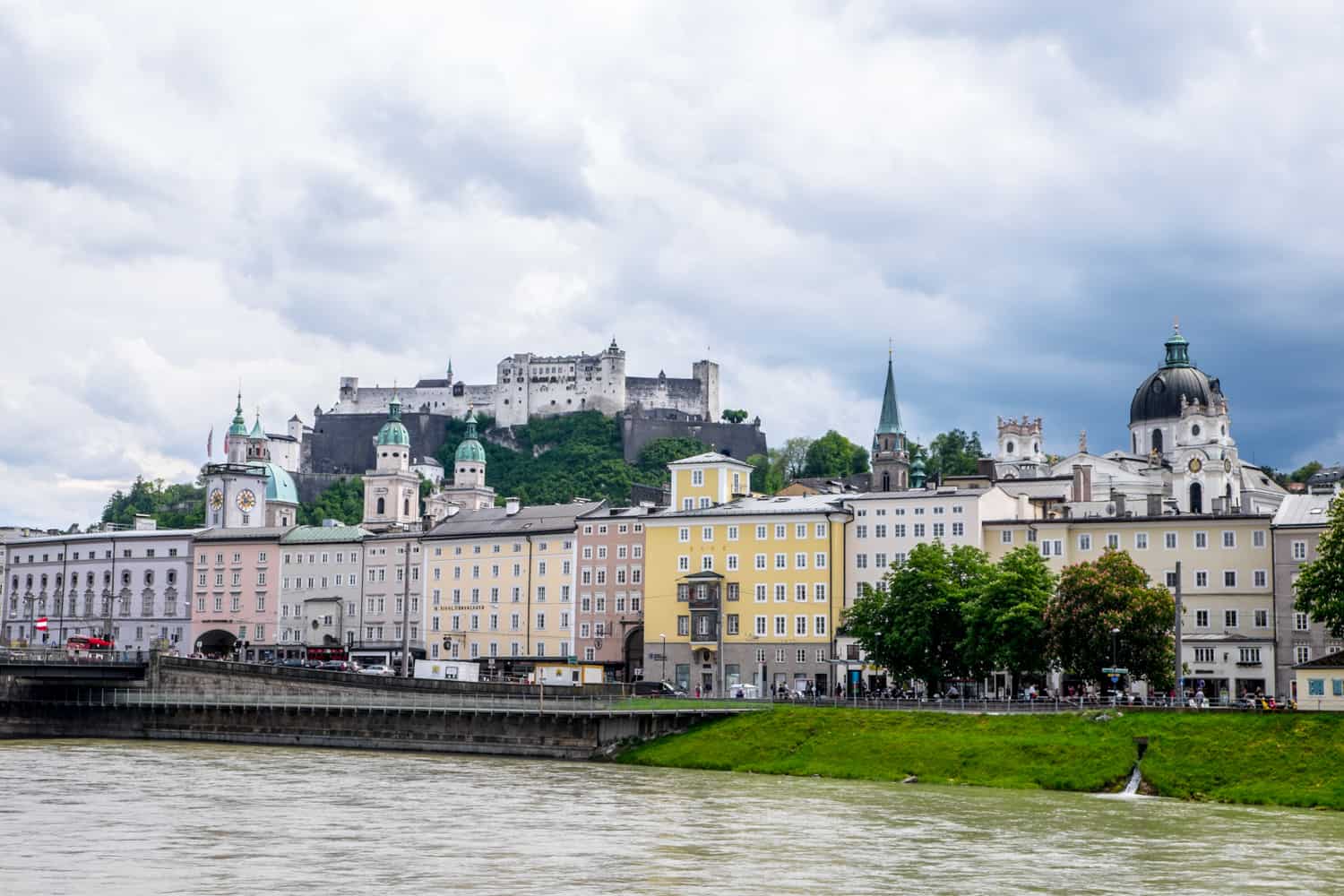 A row of rectangular buildings in pastel colours, spires and towers with mint green roofs and a castle complex on a hill - a riverside view of the UNESCO World Heritage Site of Salzburg city.