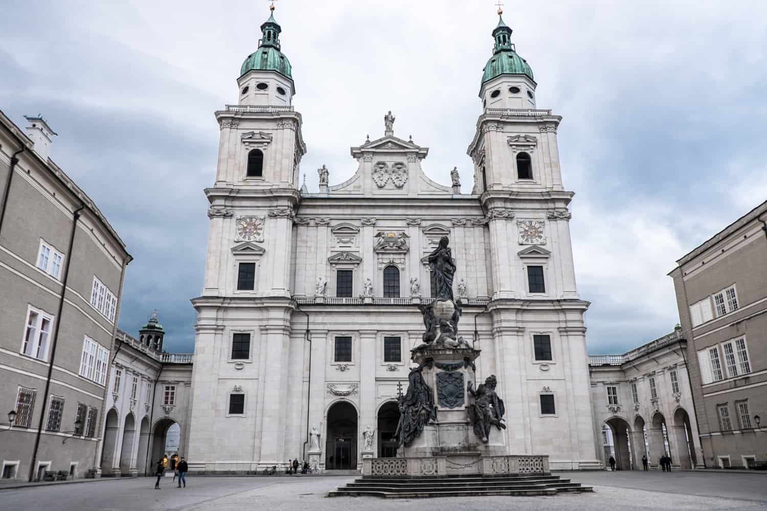 A white building with two towers with mint green spires, dominates a Public Square with a statued fountain in Salzburg, Austria.