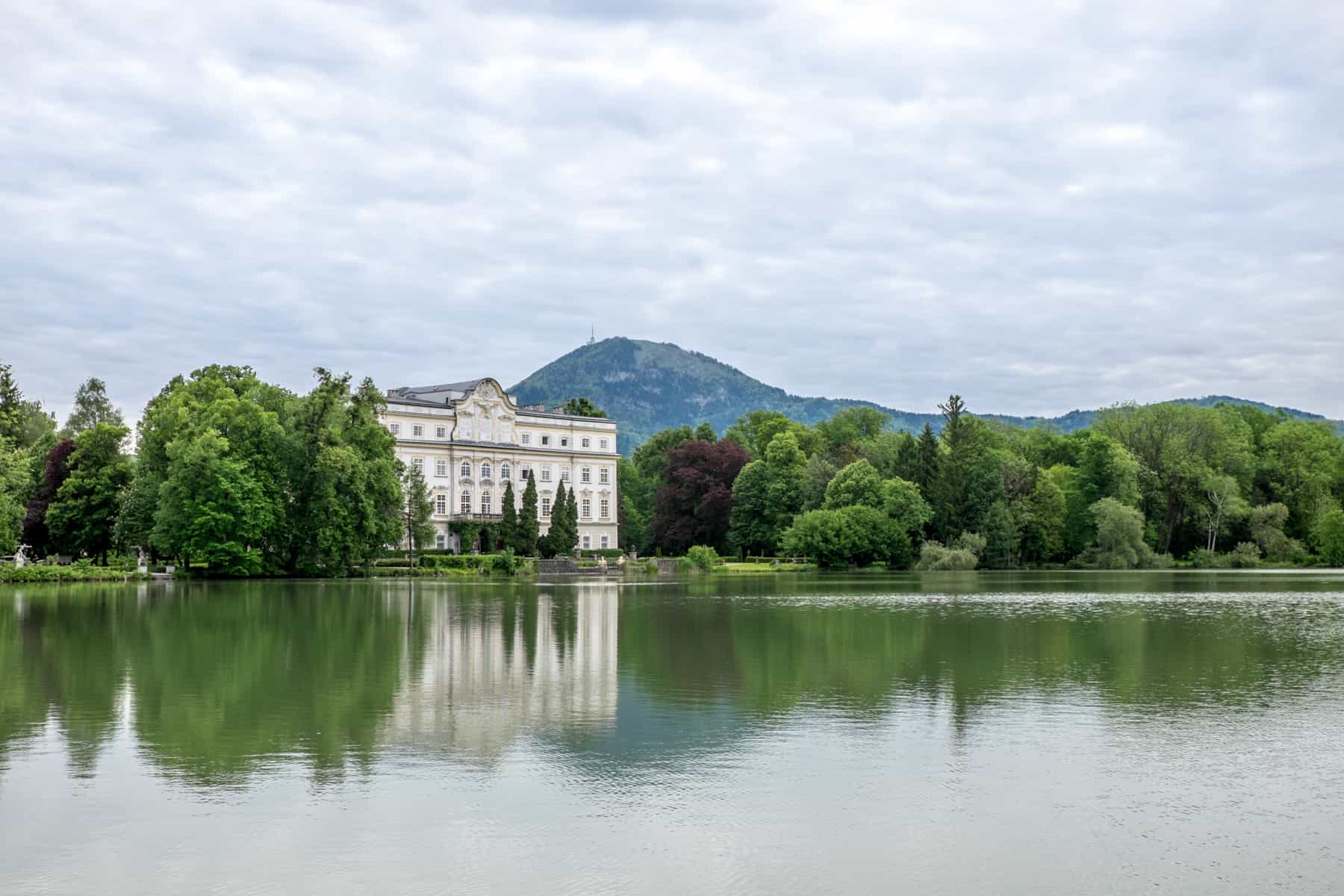 A white mansion house in Salzburg in Austria, pokes between a mass of trees and sits facing huge body of water. 