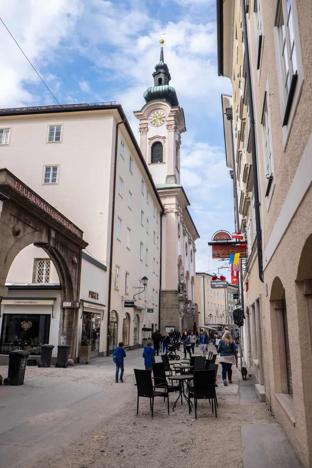 View down a long street with a brown archway and a baby pink building and clock tower with a mint green spire. Black tables and chairs can be seen in the street. 