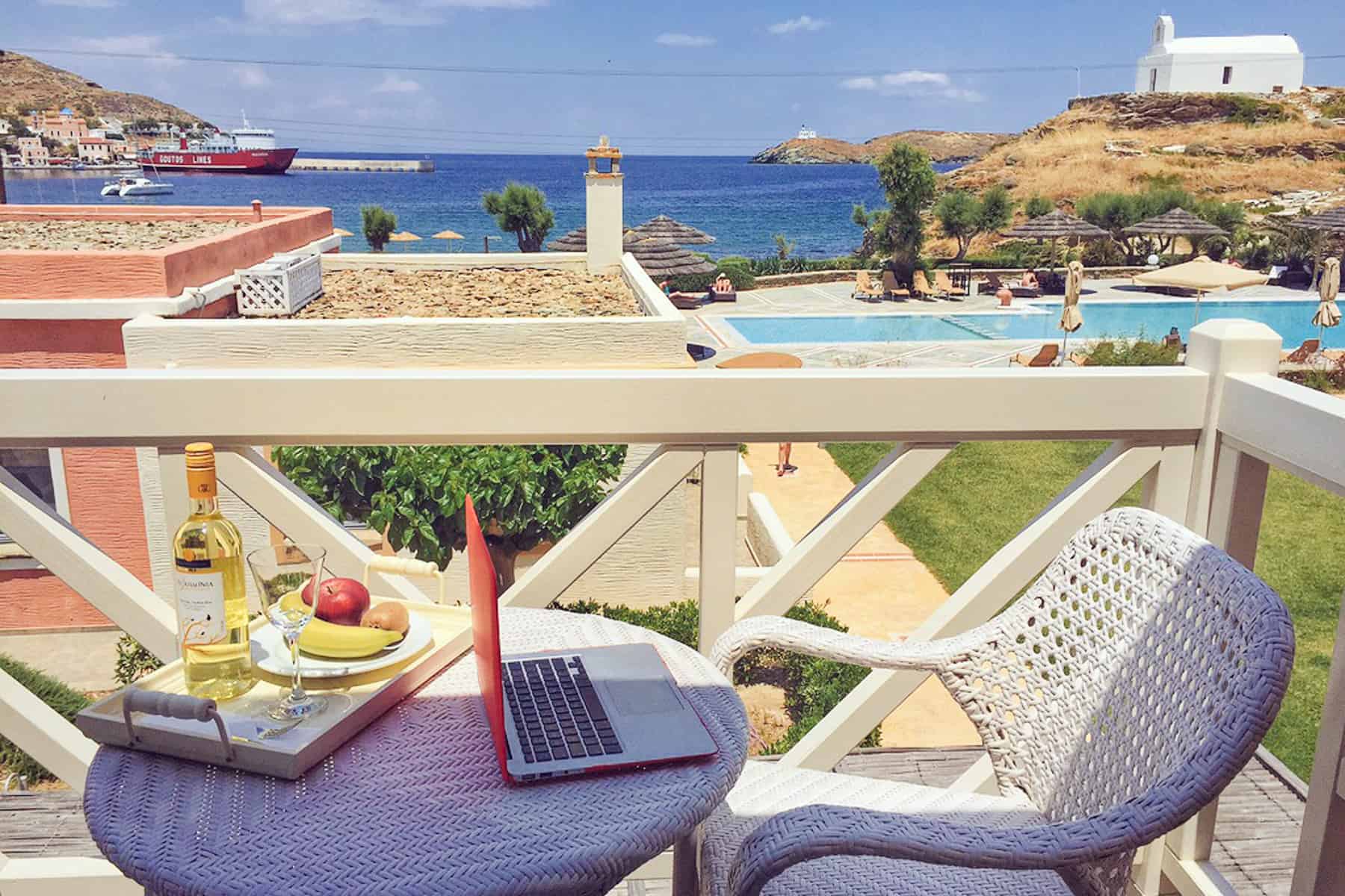 A hotel balcony view to the pool at Porto Kea Suites on Kea Island Greece. On the table is a red laptop and a bottle of white wine