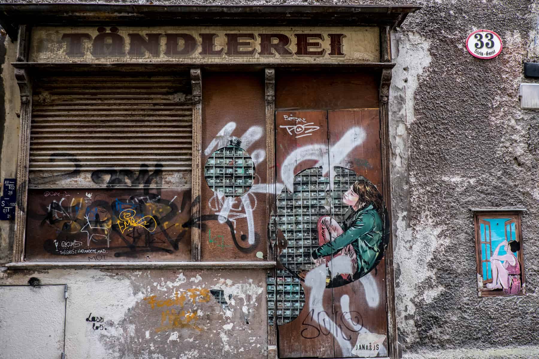 An old shop front with brown shutters, a brown door and a yellow and brown shop sign that reads "Tändlerei" is covered in modern street art paintings of women sitting in doorways and windows. 