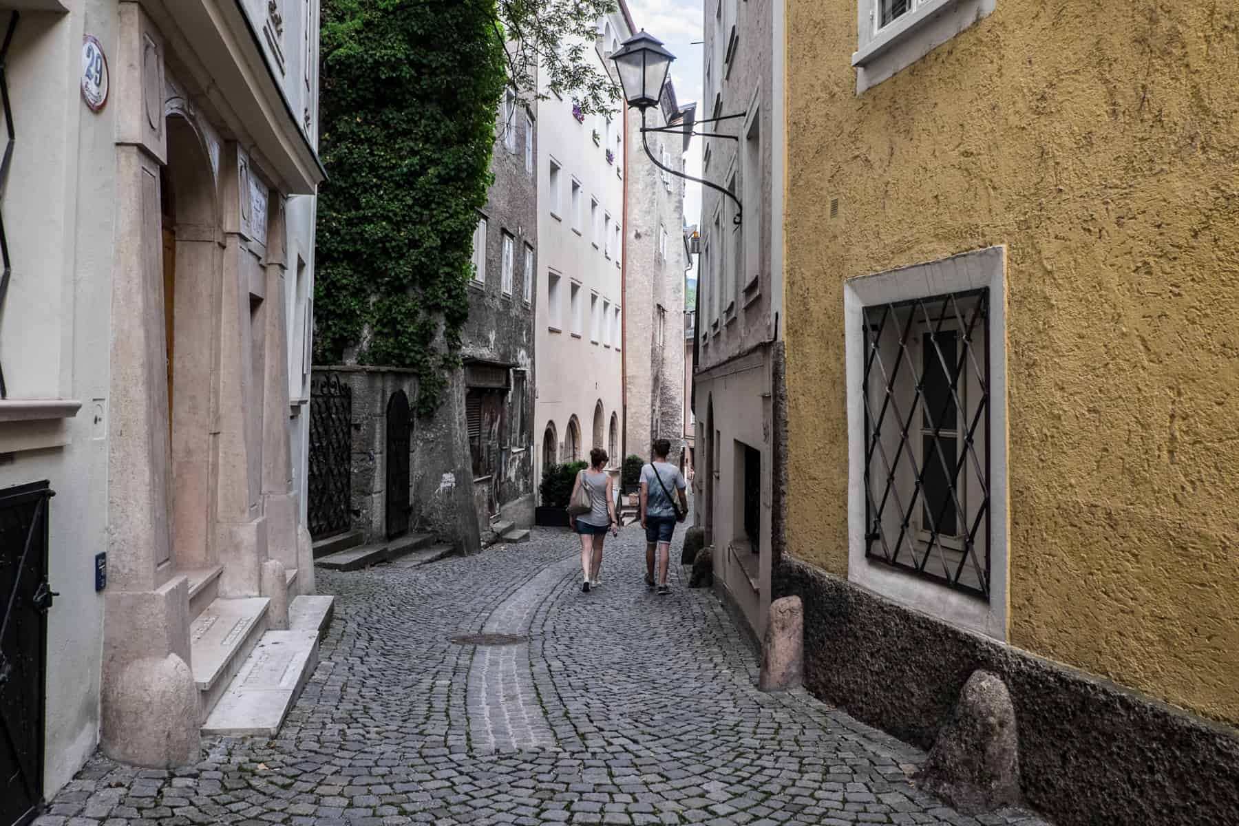 Two people walk down the oldest street a Salzburg – a wide cobblestoned street lined either side with light pink and yellow building with metal window bars and street lamps.