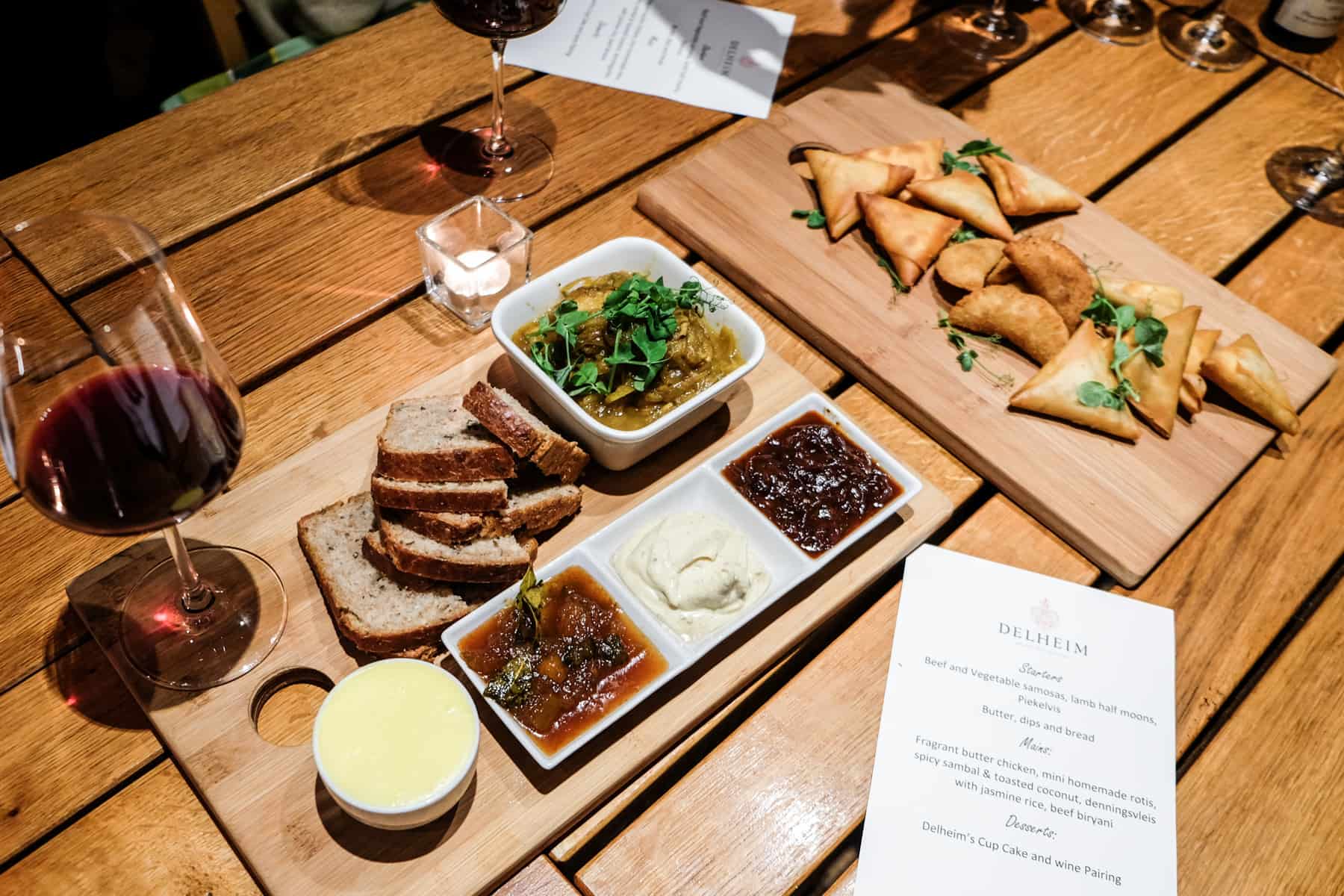 A wooden table with a menu display f a glass of red wine, bread with three different dips and samosas. A white cards displays the written menu of the Delheim Wine Estate 