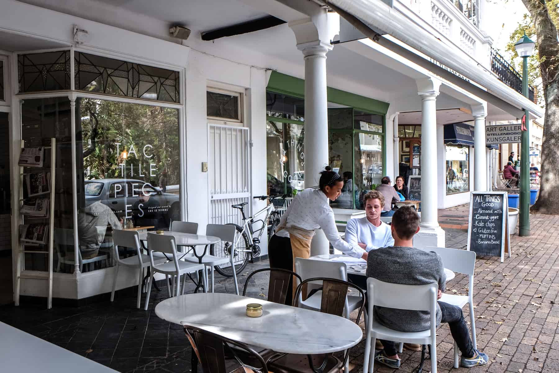 A woman serves two men at a cafe in Stellenbosch. The buildings, columns, table and chairs are all a bright white