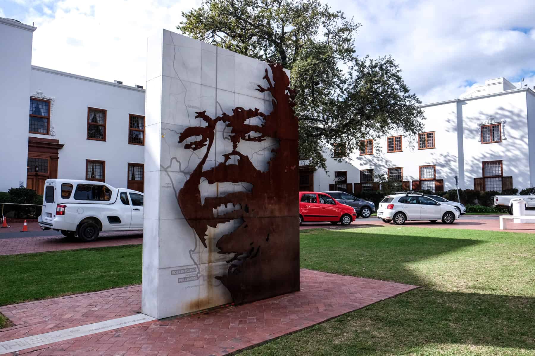 The white and brown metal art installation of the face of Nelson Mandella in Stellenbosch, set within a a green greassed courtyard of white buildings