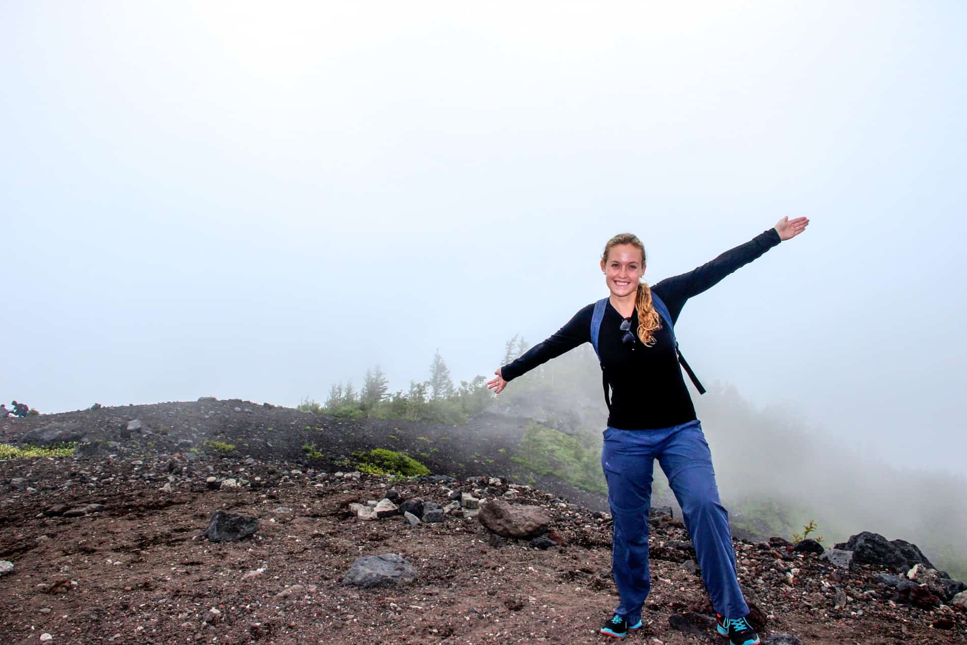 A hiker with her arms stretched, standing on a rocky mountain path with a background of fog. 