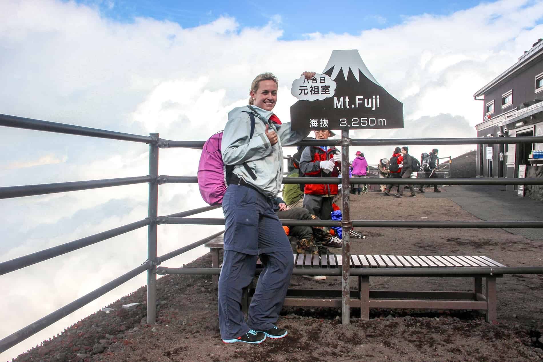 An exhausted hiker leaning on a sign at a mountain ledge which reads: Mt. Fuji 3,250m. 