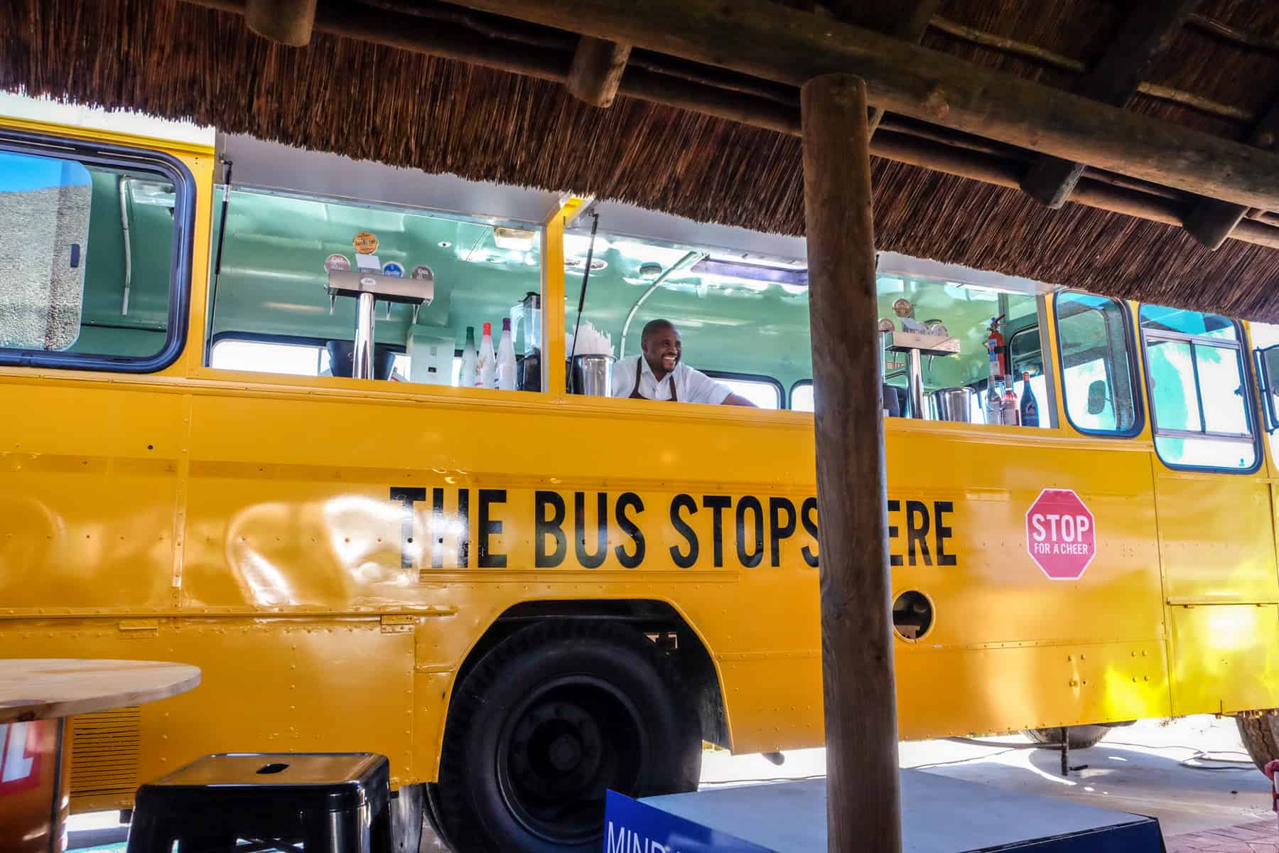 A smiling man in a white shirt stands at the counter of a refurbished yellow bus - part of the Bus Stop Cafe at Stellenbosch Vineyards 