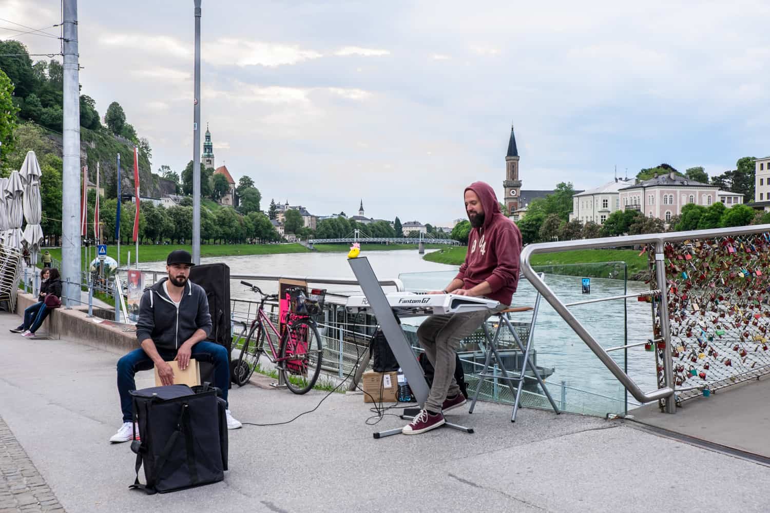 Two bearded men in hoodies - one playing a wood board drum and the other playing a white keyboard, busk next to the padlock covered river bridge in Salzburg. 
