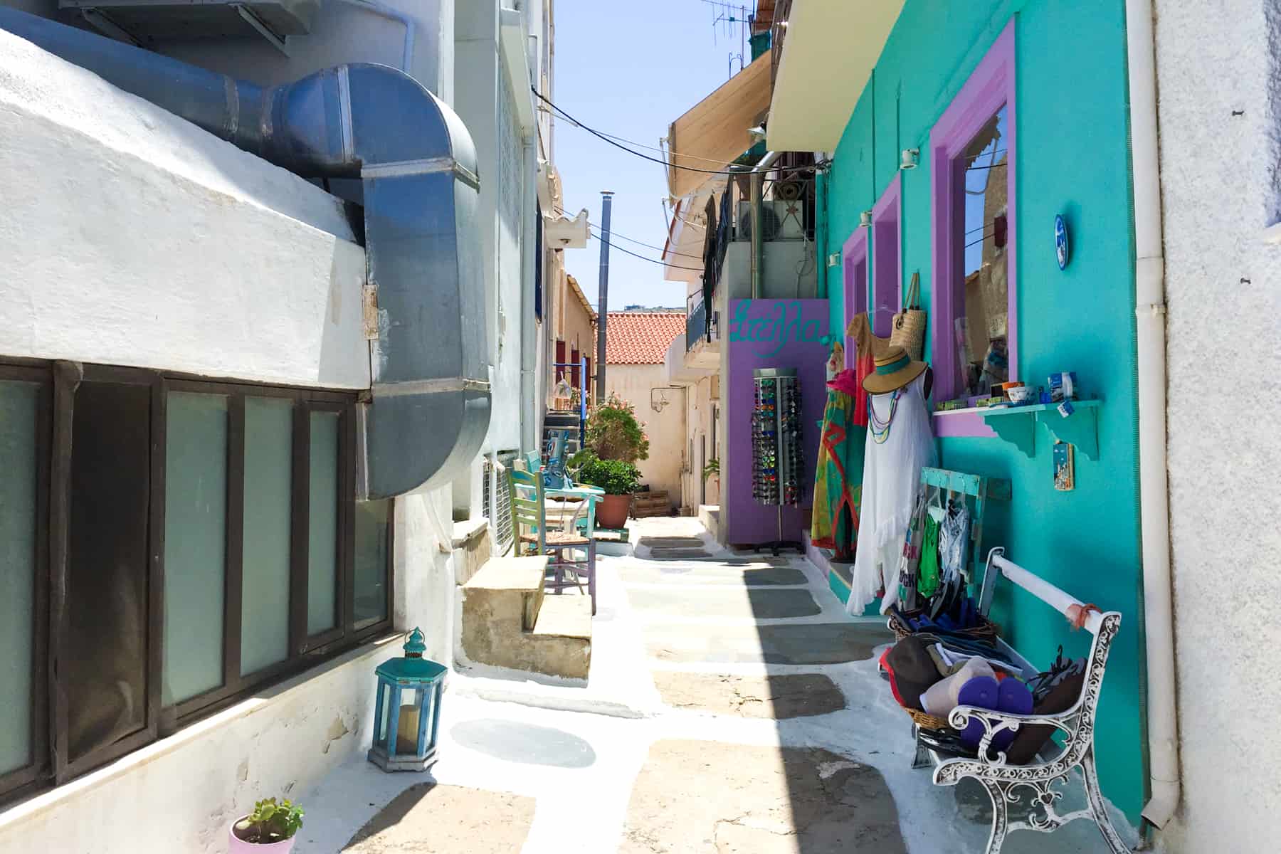 A white and stone walkway in a narrow Greek Island street lined with small white buildings and one wall painted in a bright turquoise