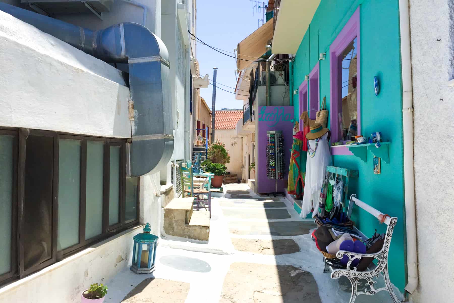 A white and stone walkway in a narrow Greek Island street lined with small white buildings and one wall painted in a bright turquoise 