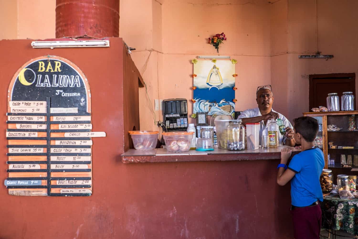 A man serves a young boy in a ochre orange painted snack bar in Cuba. The sign reads: Bar La Luna. 