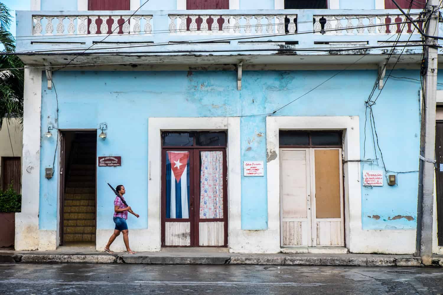 A woman holding an umbrella walked past a blue building with a Cuba flag hanging in the window. 