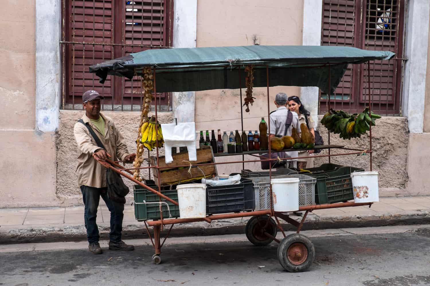 A Cuban man holds onto a wheeled metal trolley stacked with boxes, drinks bottles and bunches of bananas. 