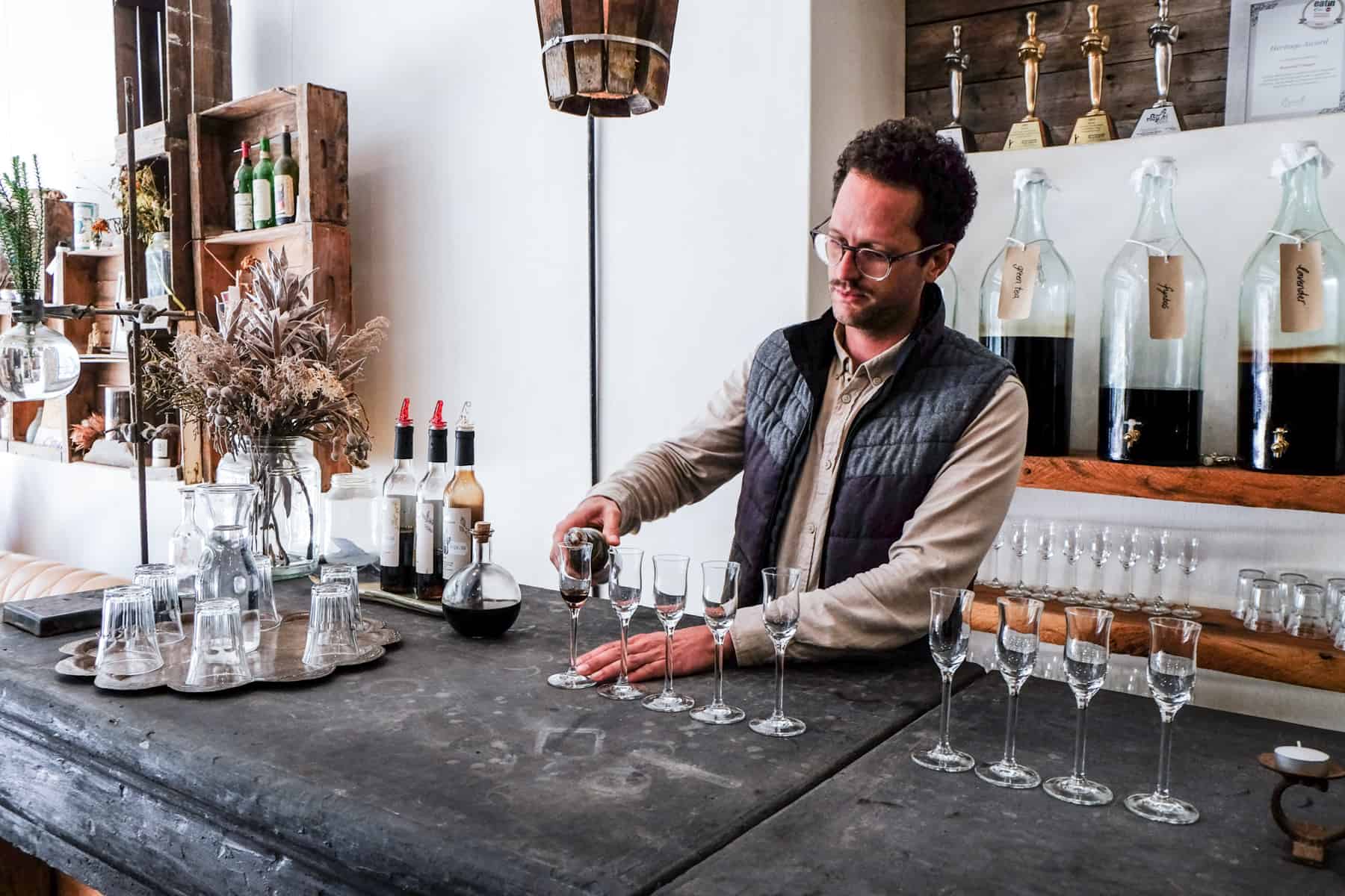 A man wearing a beige top and black waistcoat pours vinegar into tiny glasses on a dark wooden table, ready for a Vinegar tasting at Rozendal Guest Farm in Stellenbosch 